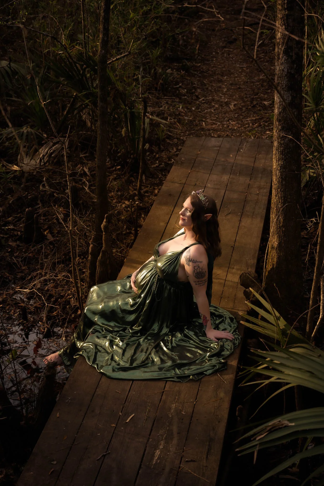 A woman dressed as an elf or fairy with pointed ears and a crown, sitting on a wooden platform in a forest, wearing a shiny green dress, surrounded by trees and foliage at dusk.