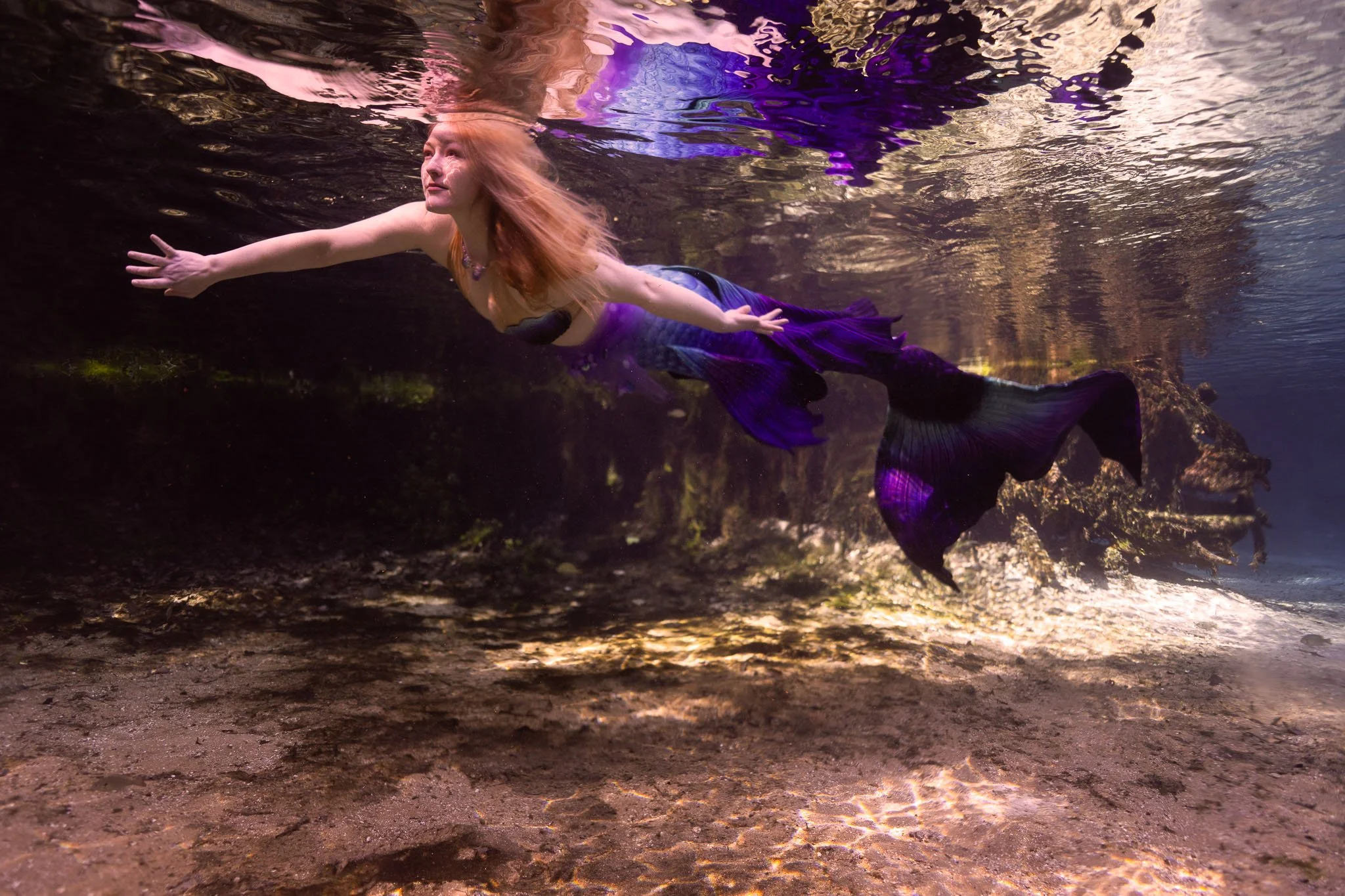 A woman with long red hair swimming underwater in a river with a purple mermaid tail, reaching out with her arms extended.