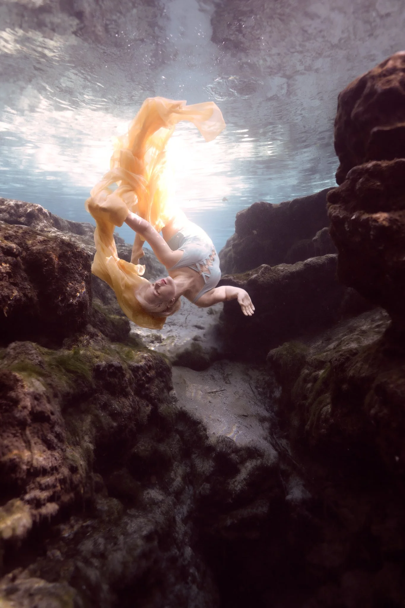 An underwater scene with rocks and a person in a pale dress lying among rocks, surrounded by clear water.