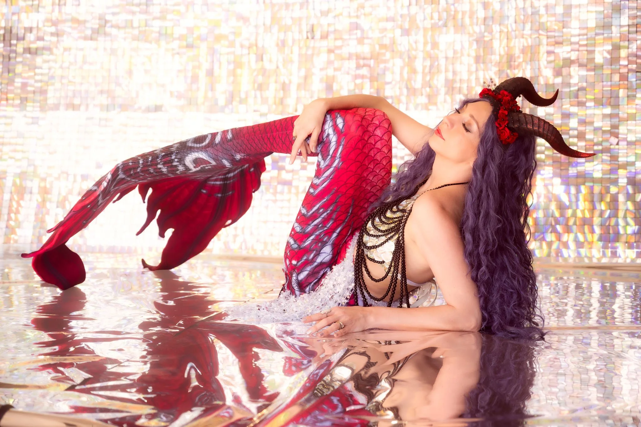 A woman with purple hair and horn-shaped headpiece, dressed in a fantasy costume, is reclining in shallow water with a colorful, reflective backdrop.