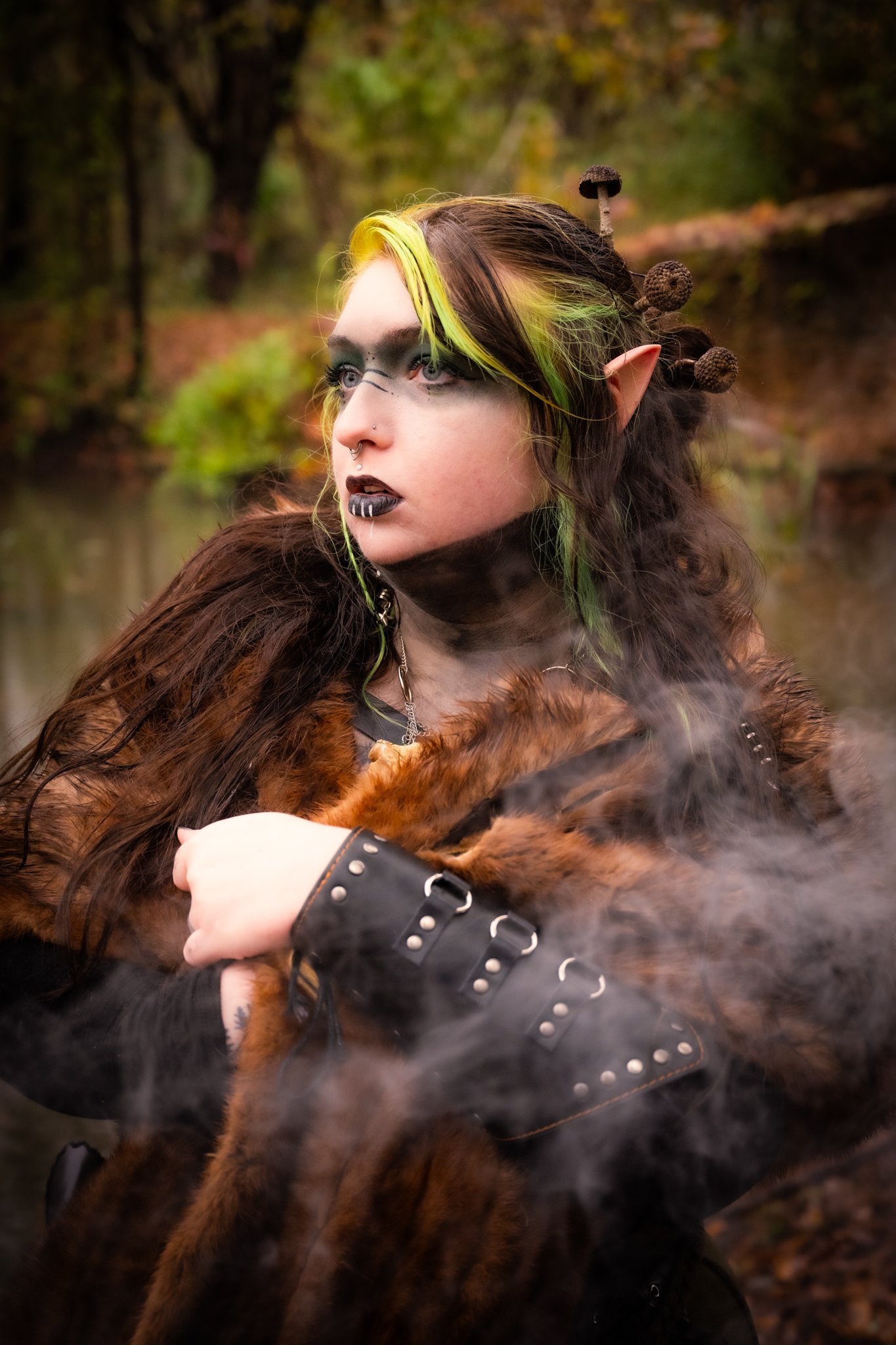 Person with fantasy makeup and elf ears, wearing a faux fur coat and leather wrist cuffs, outdoors with trees and fallen leaves in background.