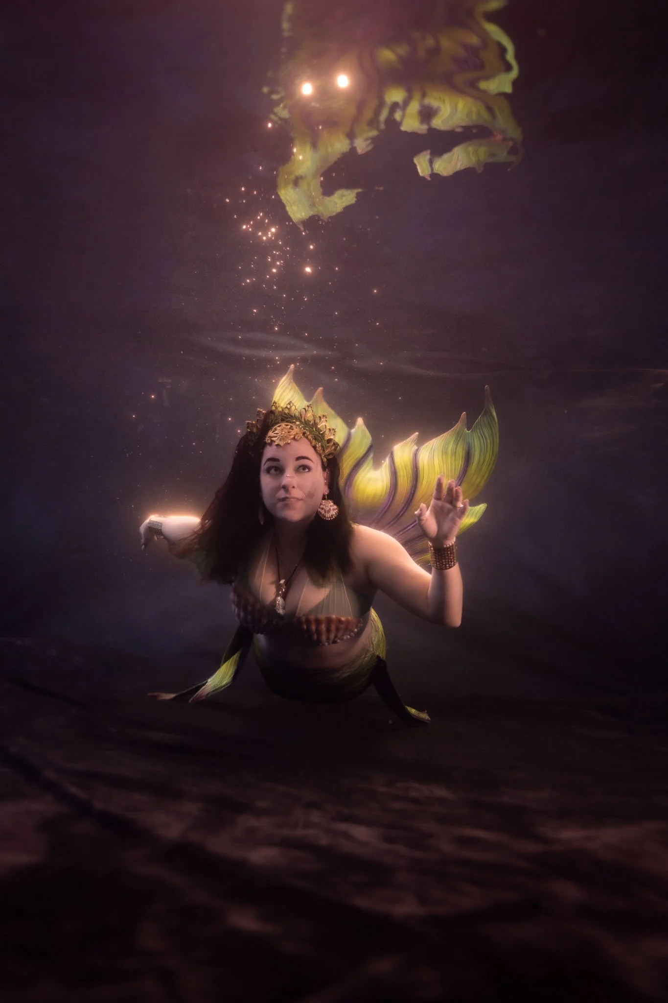 A person dressed as a mermaid with a yellow and purple tail and a decorative headpiece, swimming underwater with bubbles around them.