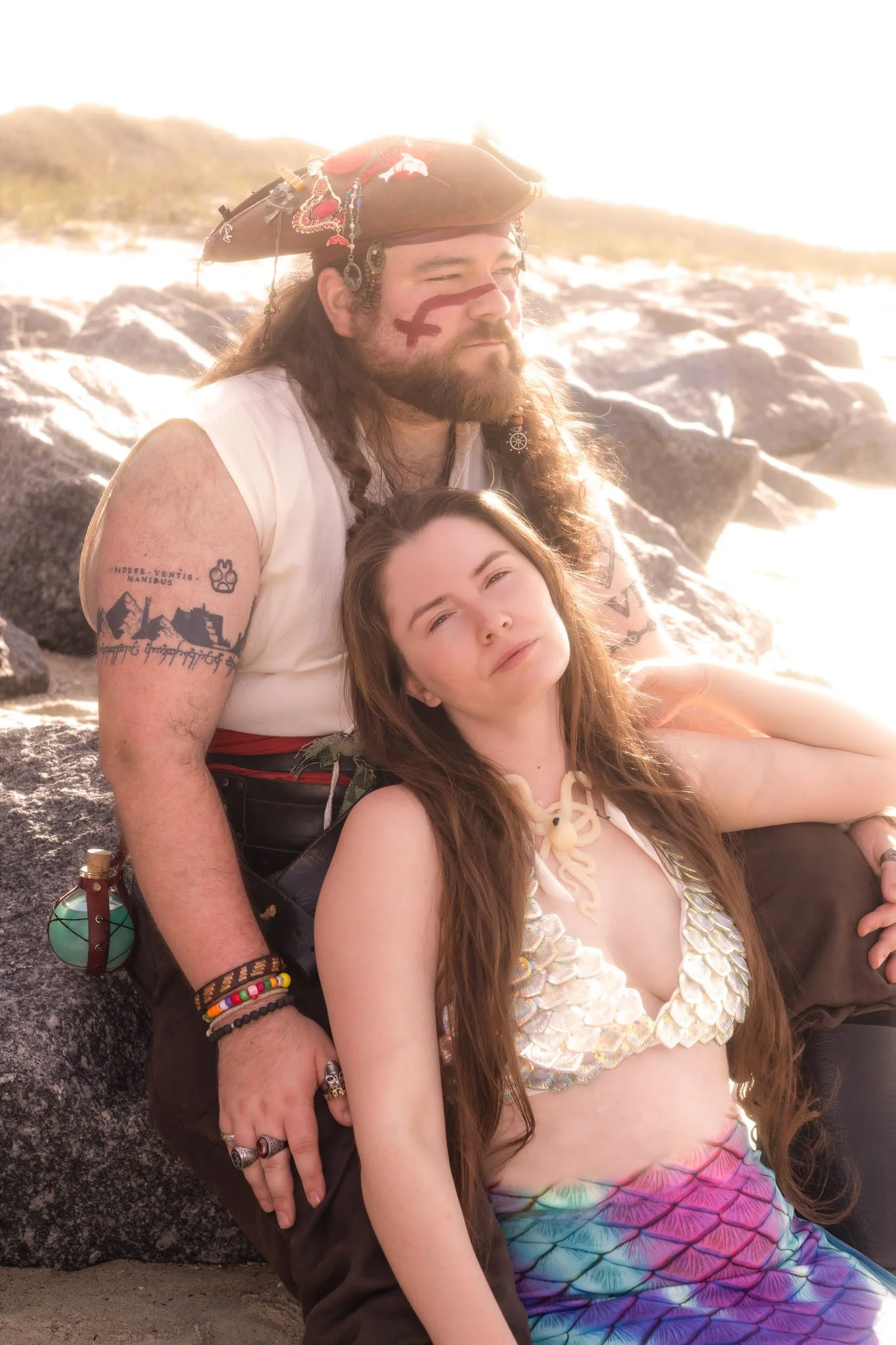 A man dressed as a pirate with a colorful hat and tattoos, sitting next to a young woman with long hair wearing a seashell costume, both relaxing on a rocky beach at sunset.