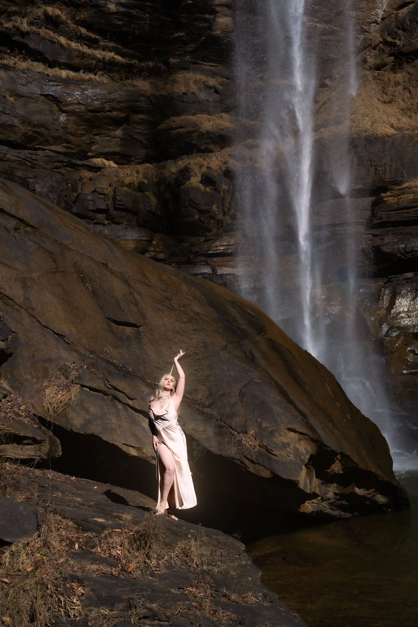 A woman in an elegant beige dress with a slit, standing on rocks near a waterfall, holding her right arm高 up, with water cascading down a rocky cliff behind her.
