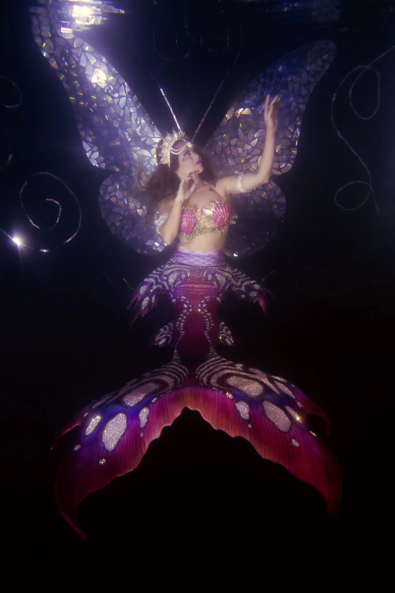 A person wearing a butterfly costume with iridescent wings, a headband, and detailed clothing, posing underwater or against a dark background with light reflections.