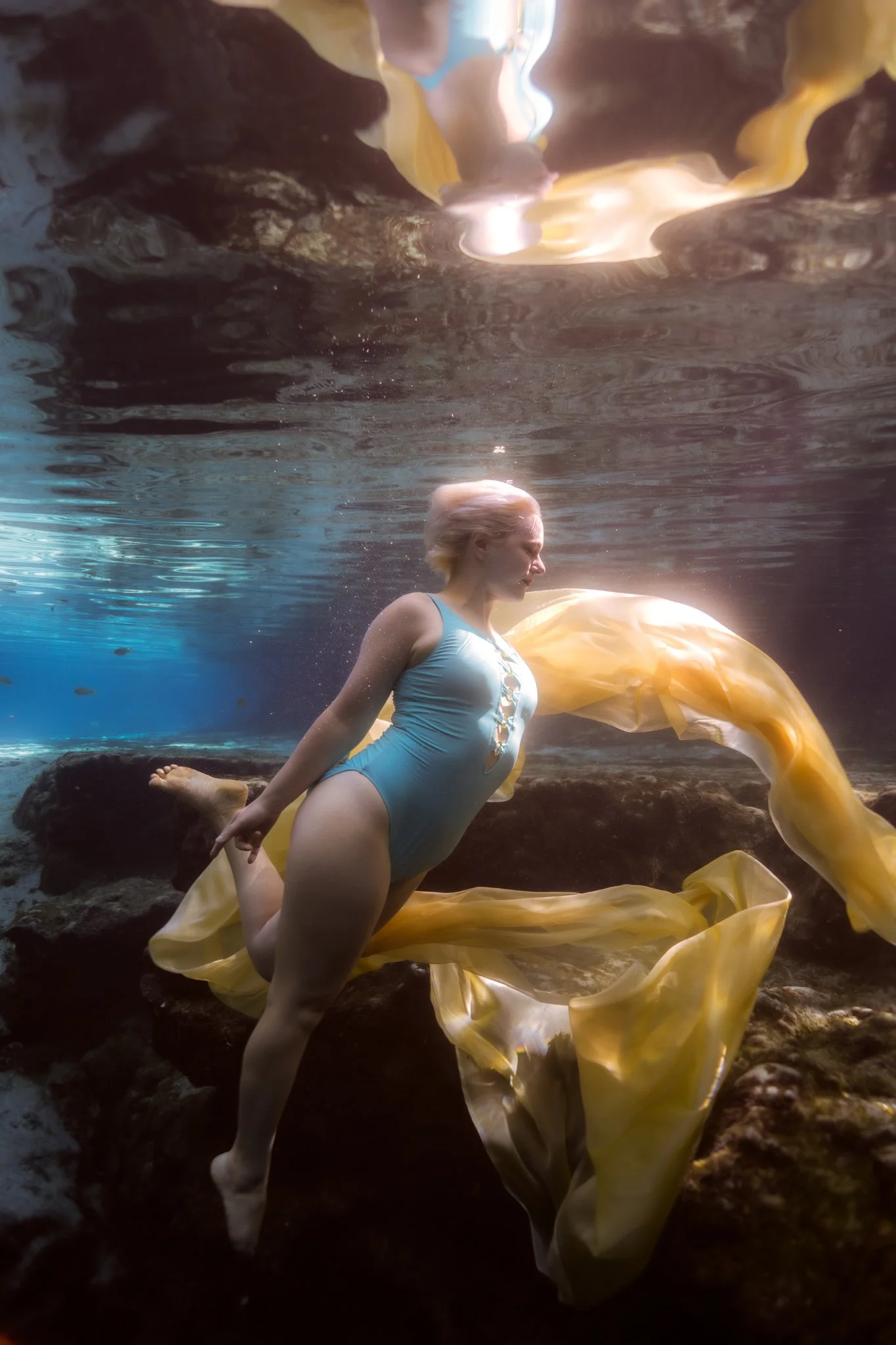 A woman with blonde hair tied back, wearing a light blue swimsuit, underwater near rocks, with flowing yellow fabric around her.