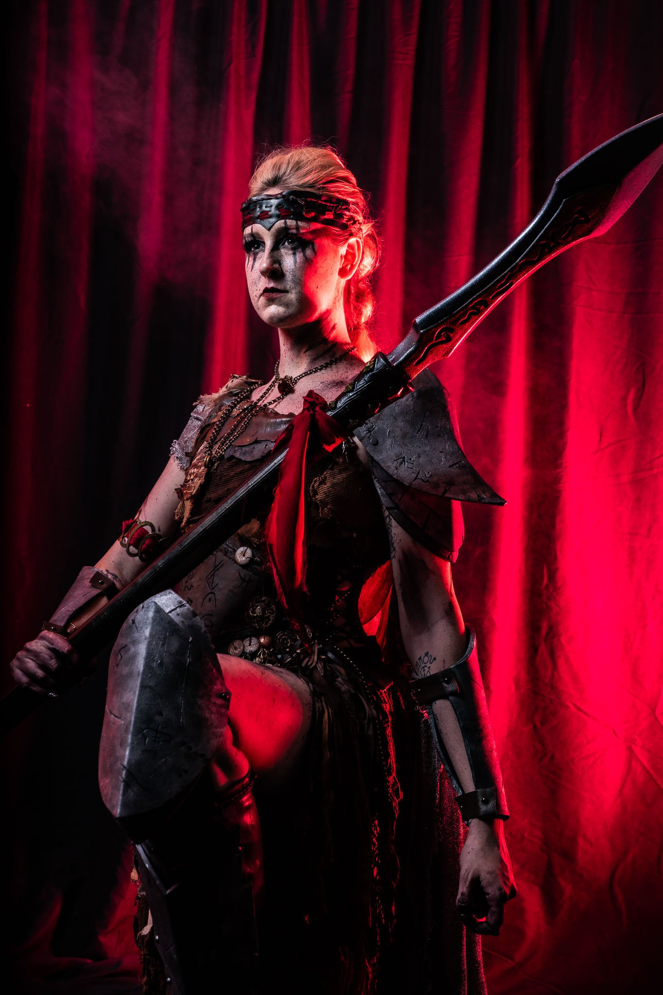 A woman with blonde hair, black makeup, and tattoos, dressed in dark clothing, sitting against red velvet curtains, holding a large black and red sword.
