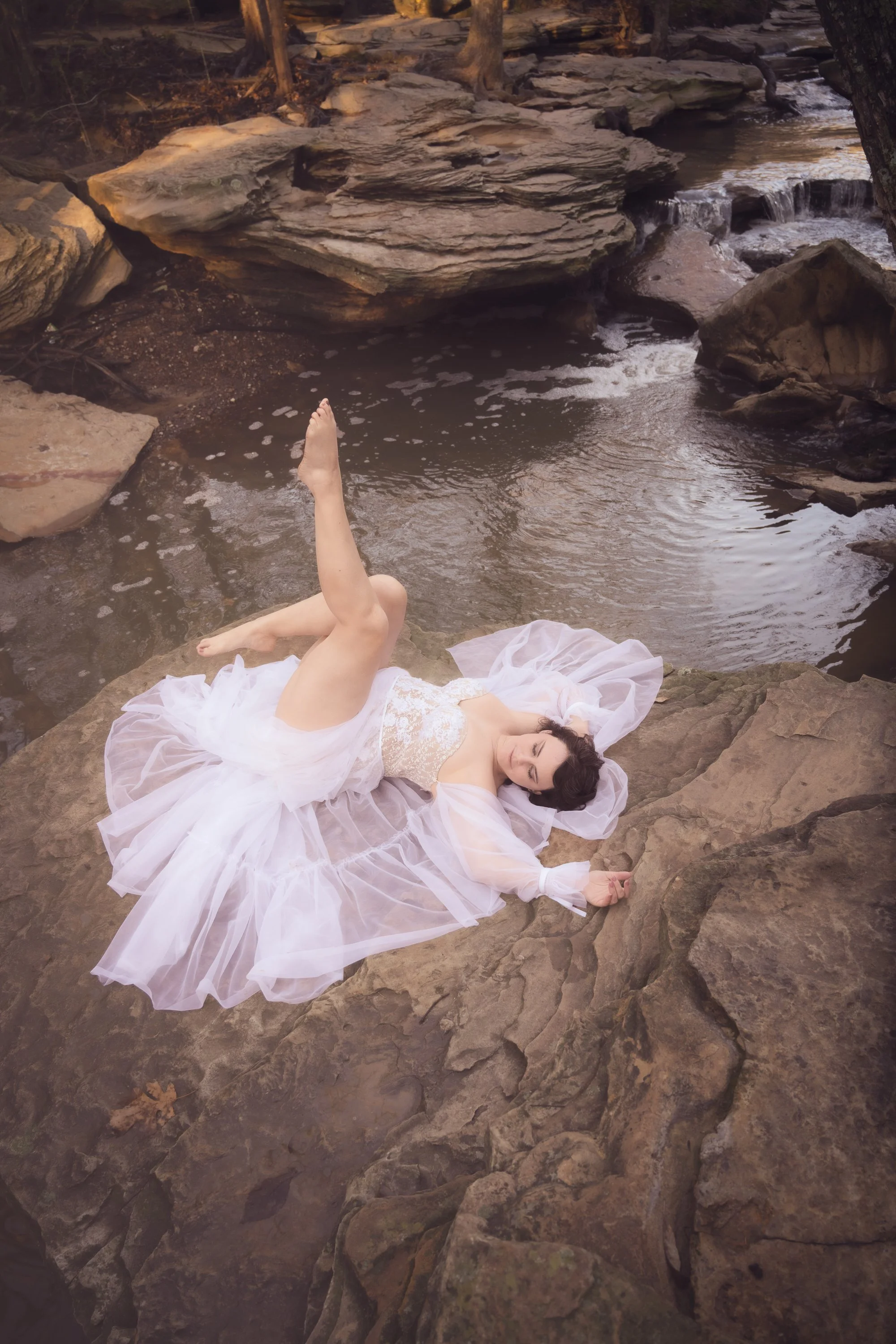 A woman dressed in a white flowing gown lies on rocks near a small waterfall and stream, surrounded by natural outdoor scenery.
