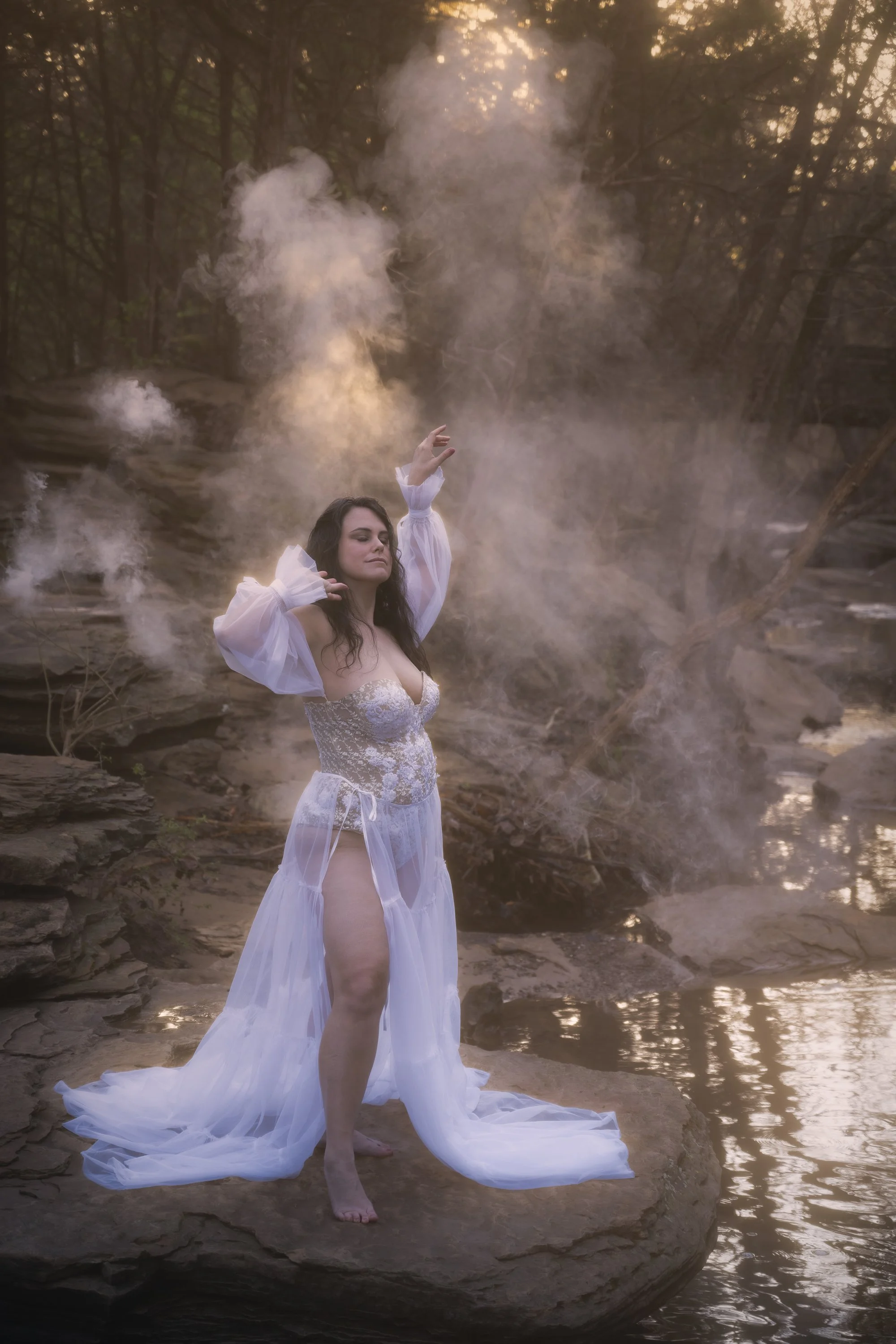 A woman dressed in white, semi-transparent clothing stands on a rocky riverbank surrounded by water, with smoke or mist rising behind her and trees in the background, during sunset or early evening.