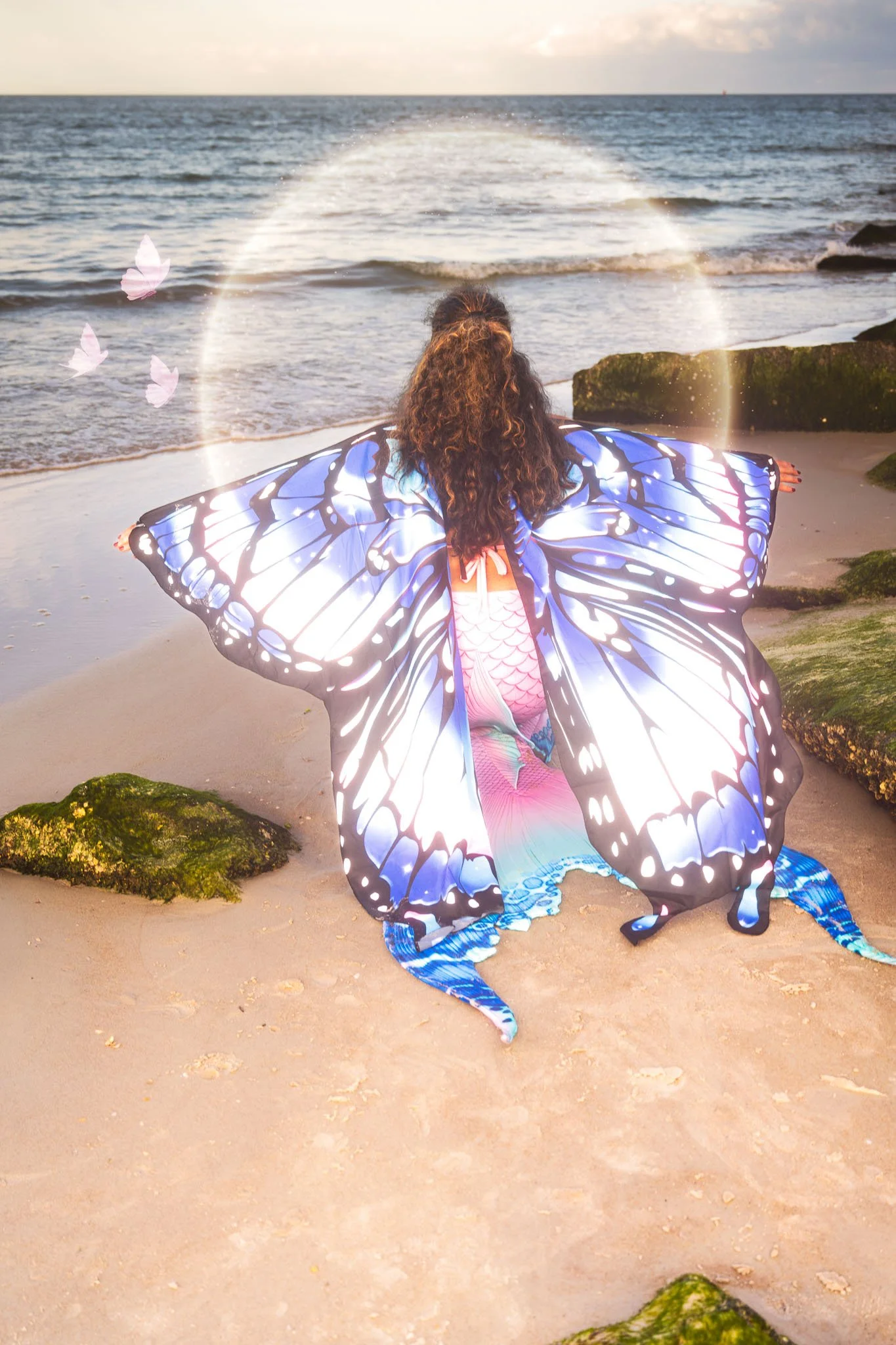 A person with long, curly hair wearing a butterfly-shaped costume with blue, black, and white patterns sitting on a sandy beach facing the ocean during sunset, with a visible rainbow halo, rocks in the foreground, and butterflies flying near the pers