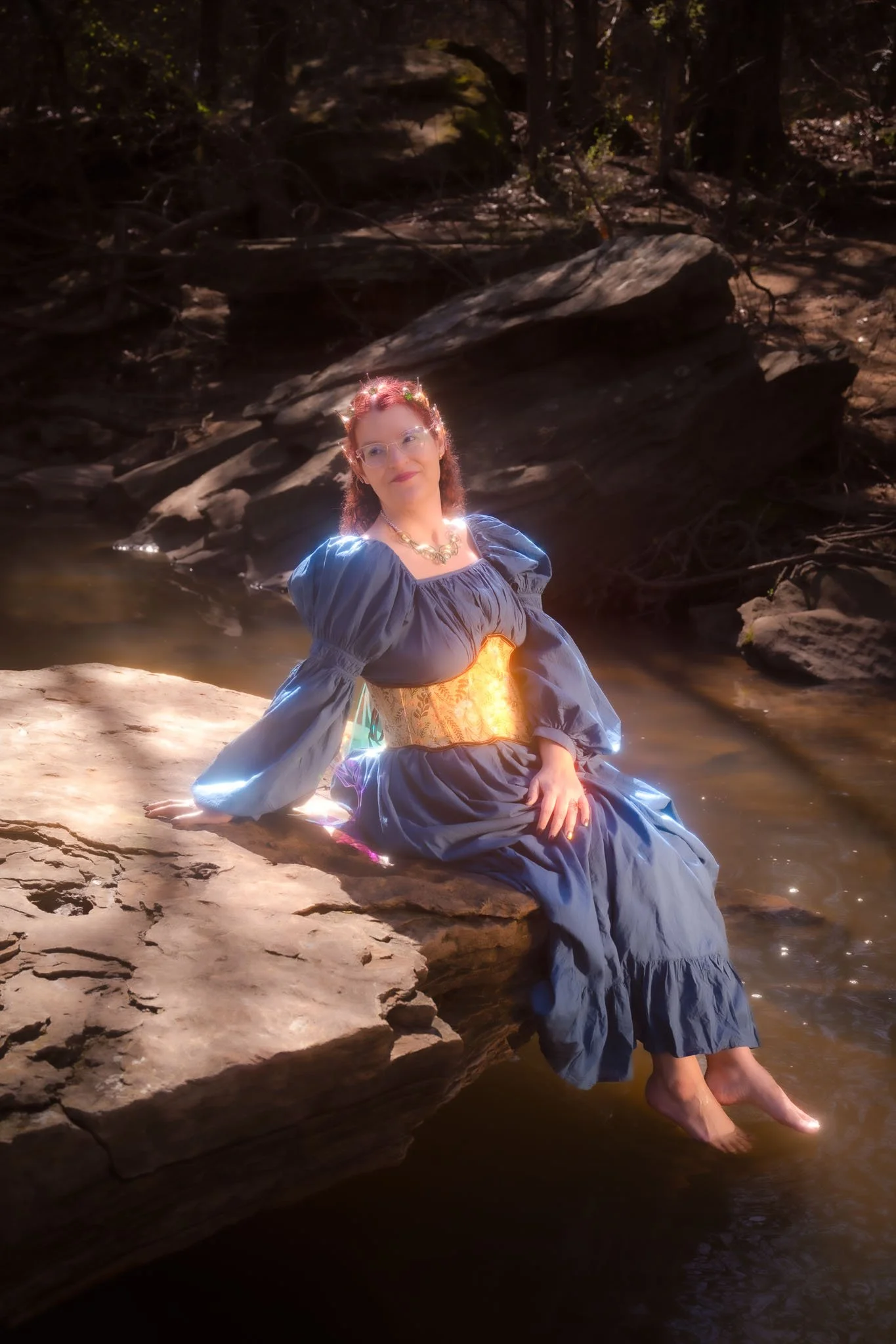 A woman in a blue dress with puffy sleeves and a gold corset sits on a rock at the edge of a river, with her feet dipped in the water. She wears glasses and a floral headband, and there is sunlight illuminating her and reflecting off the water.