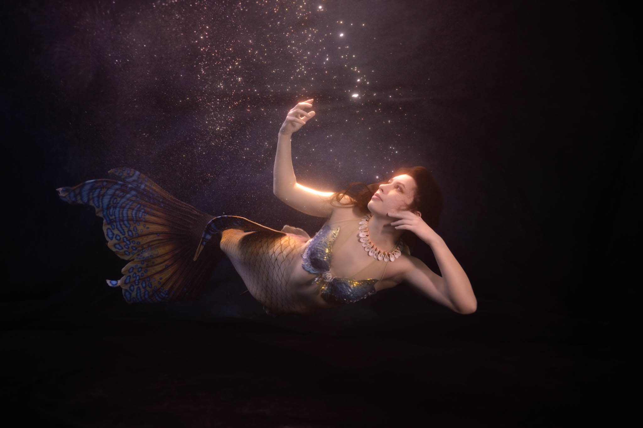 A woman dressed as a mermaid with a scaled tail, lying underwater and reaching towards floating particles or bubbles with her right hand, while her left hand touches her face, illuminated by soft light.
