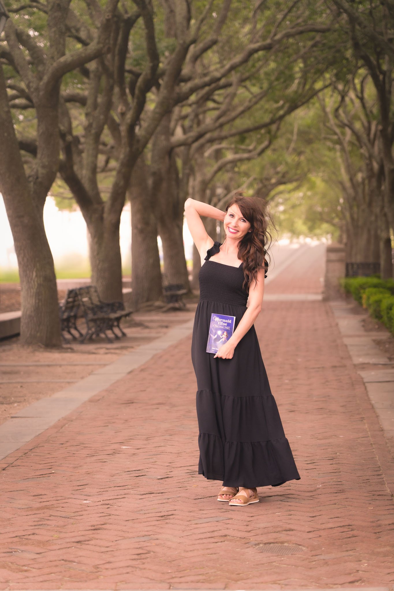 A woman in a black dress and beige sandals standing on a brick-paved walkway in a park with trees along the sides. She is smiling and holding a purple brochure about 'The Little Mermaid' movie.