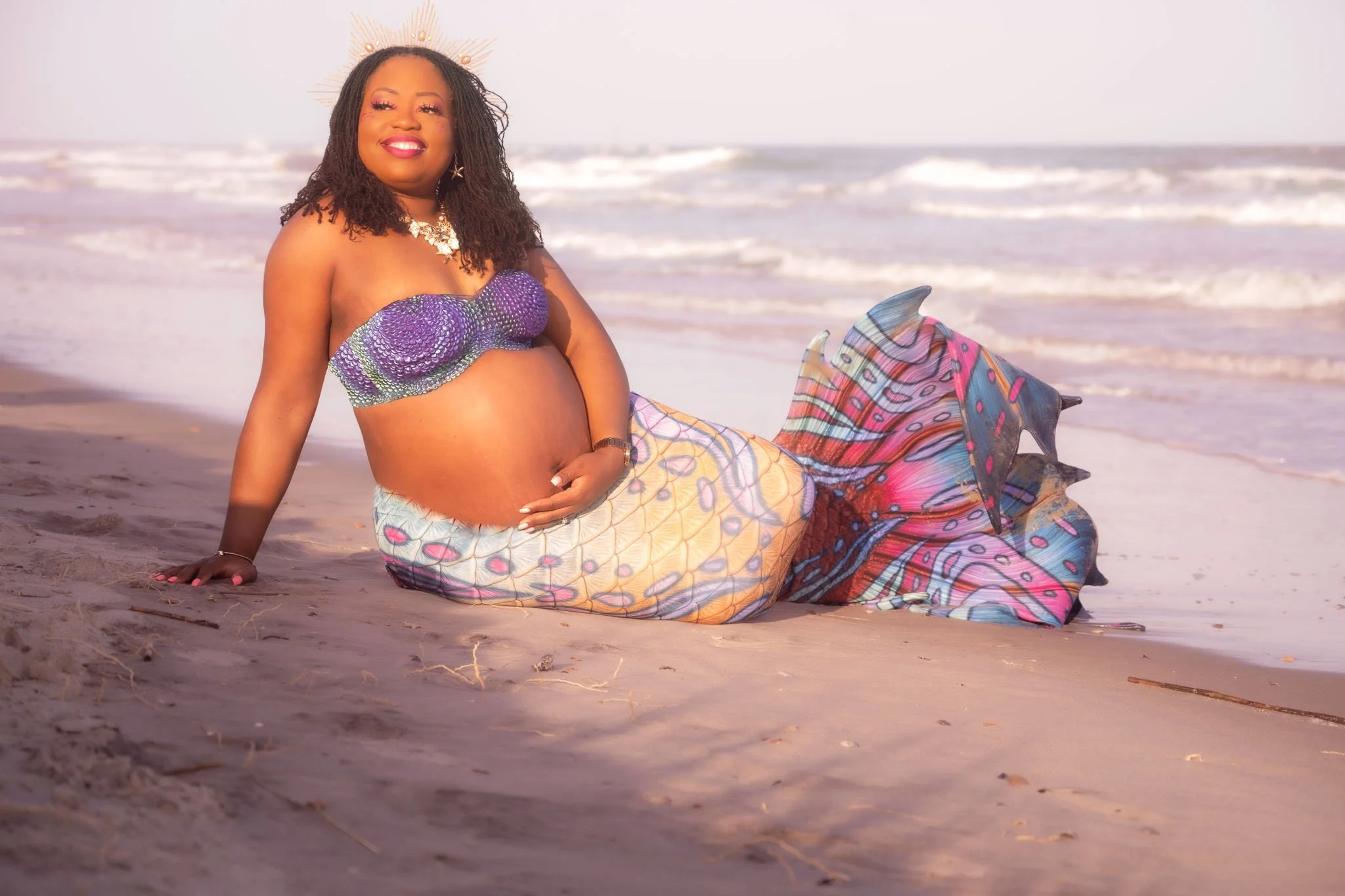 A woman with a mermaid tail sitting on the beach near the shoreline, wearing a purple seashell bikini top, smiling, with the ocean in the background.
