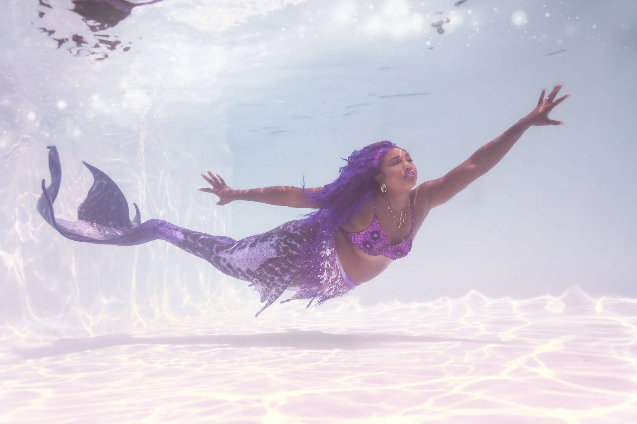 A woman with purple hair and a purple mermaid costume swimming underwater in a pool, reaching with her right hand.