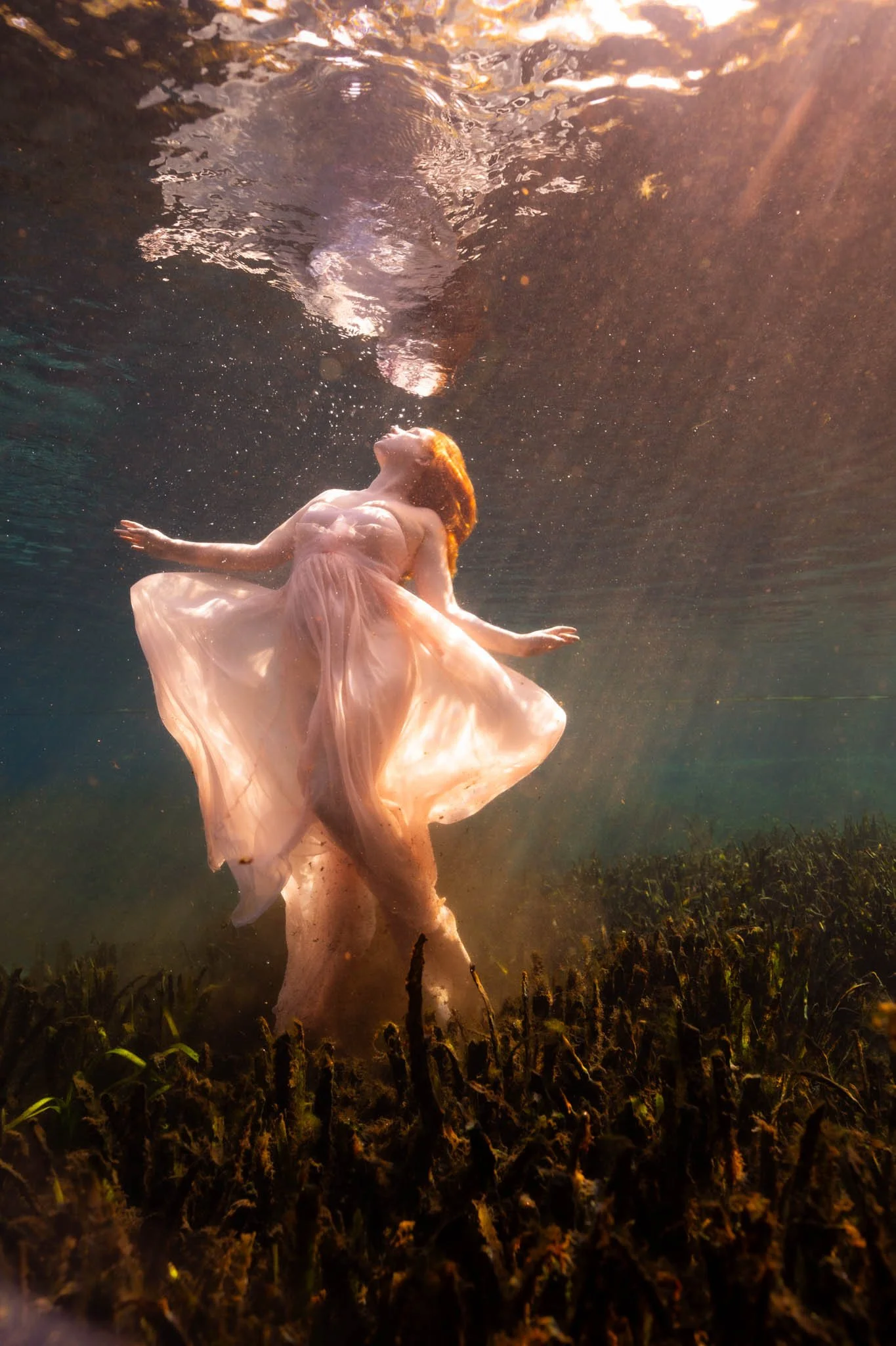 A woman in a flowing pink dress swimming underwater with sunlight filtering through the water surface.