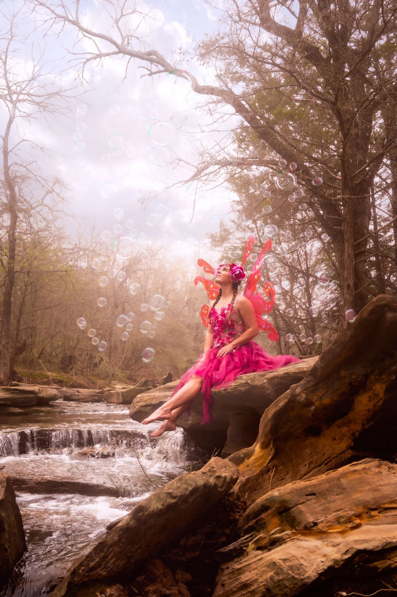 A woman dressed as a fairy with pink wings and a pink dress sits on rocks by a stream in a forest, with trees and cloudy sky above and bubbles floating around.