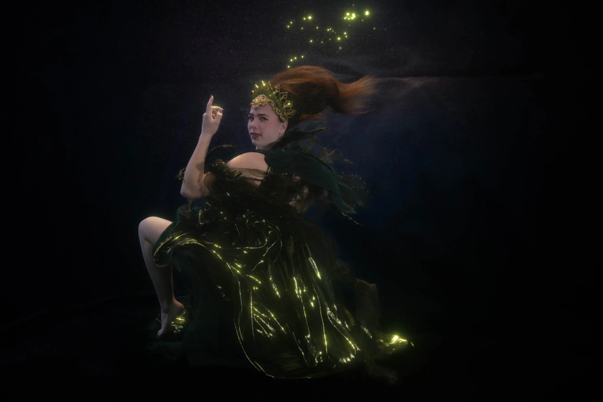 A woman with long hair floating underwater, wearing a dark dress with glowing yellow patterns and a gold headpiece with green leaves. Bright yellow bubbles or light particles surround her.