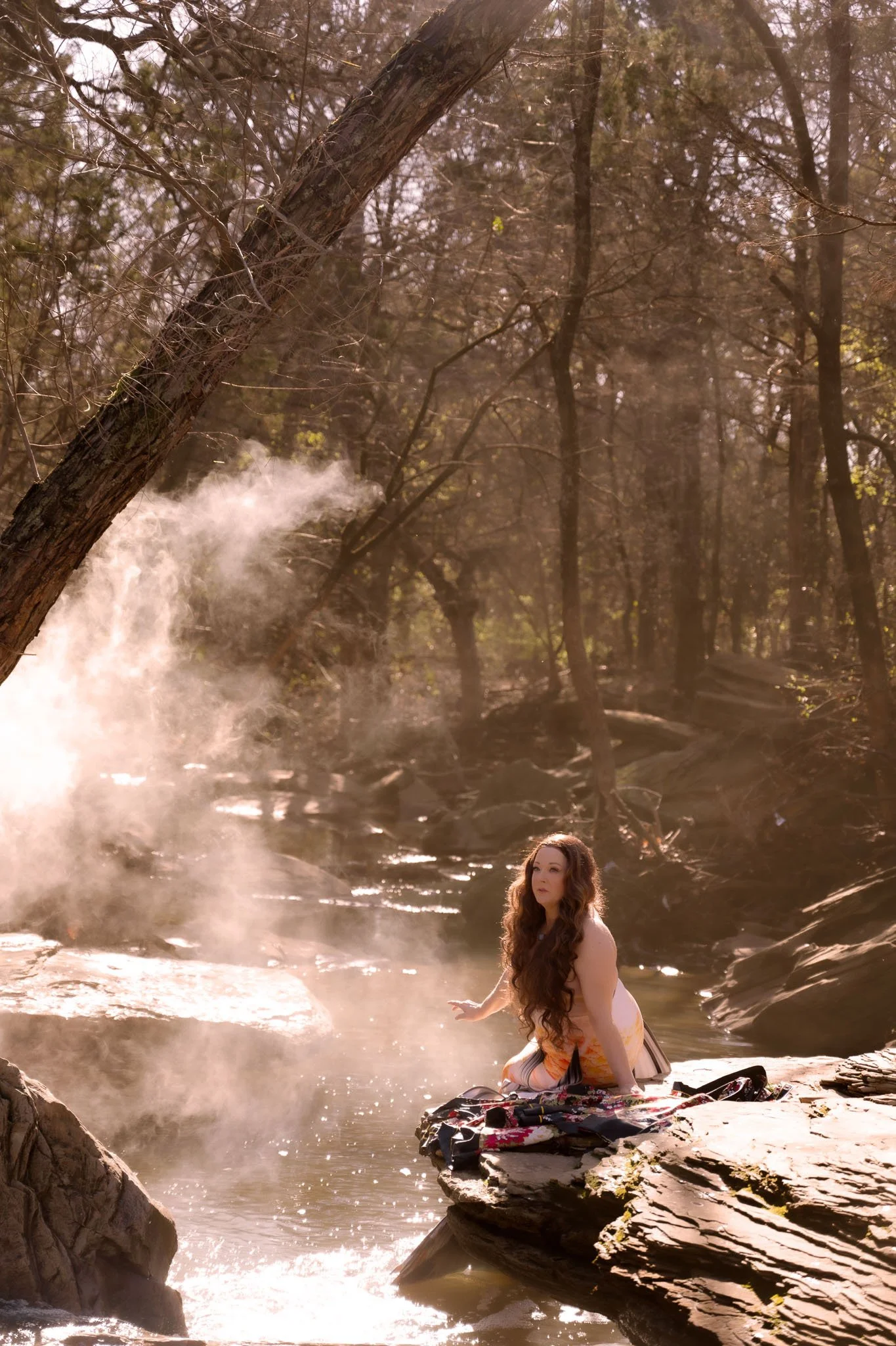 A woman with long, curly hair wearing a white dress sits on a rock by a steaming hot spring or river in a forest with tall trees and sunlight filtering through the branches.