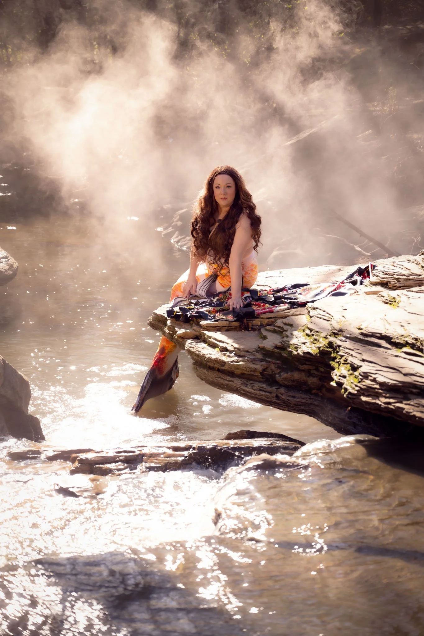 A woman with long, wavy brown hair sitting on a large rock near a steam-filled hot spring or geyser, with mist rising behind her and water flowing around the rocks.