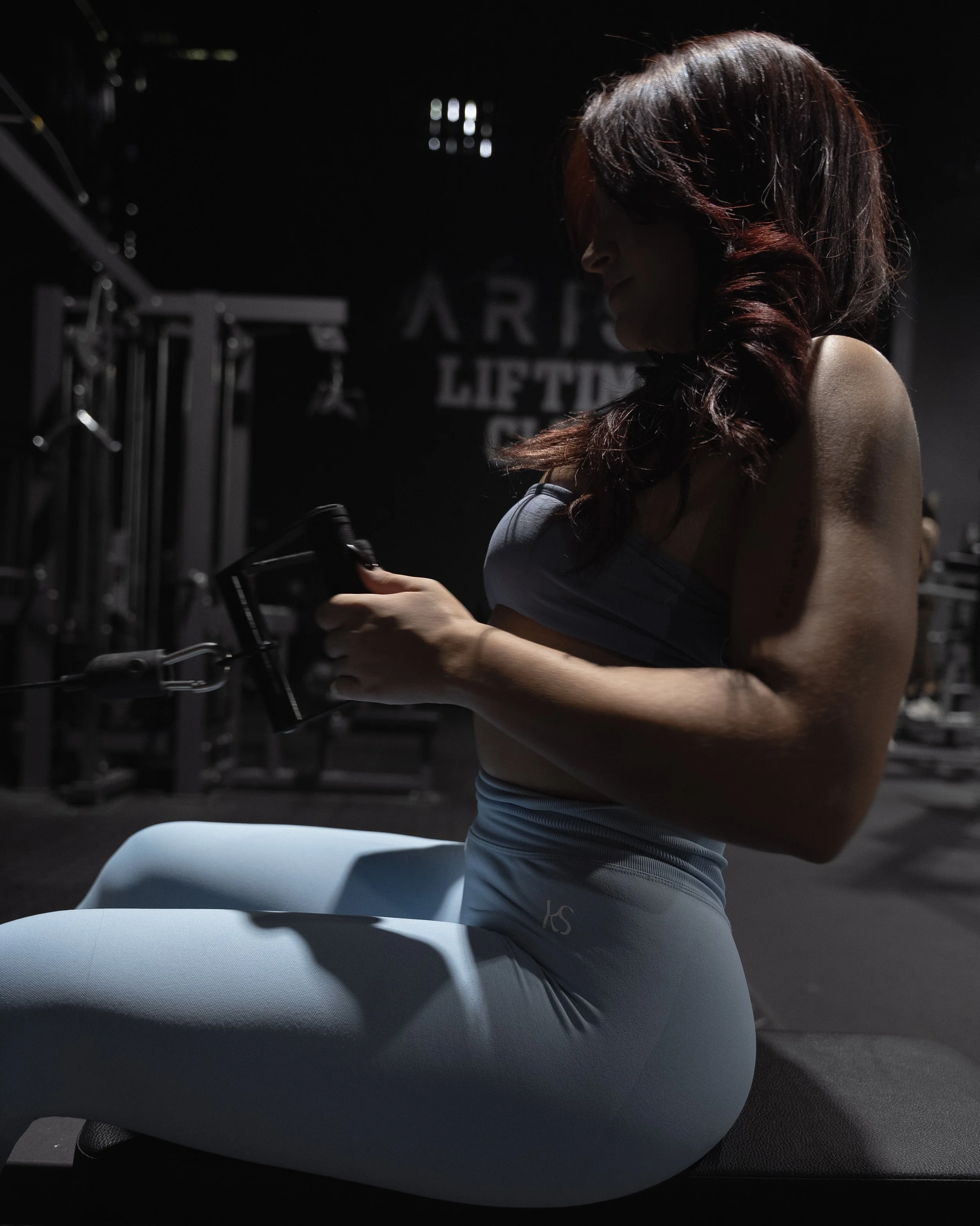 Woman with red hair in workout attire sitting on a light blue exercise ball in a dark gym, holding a dumbbell.