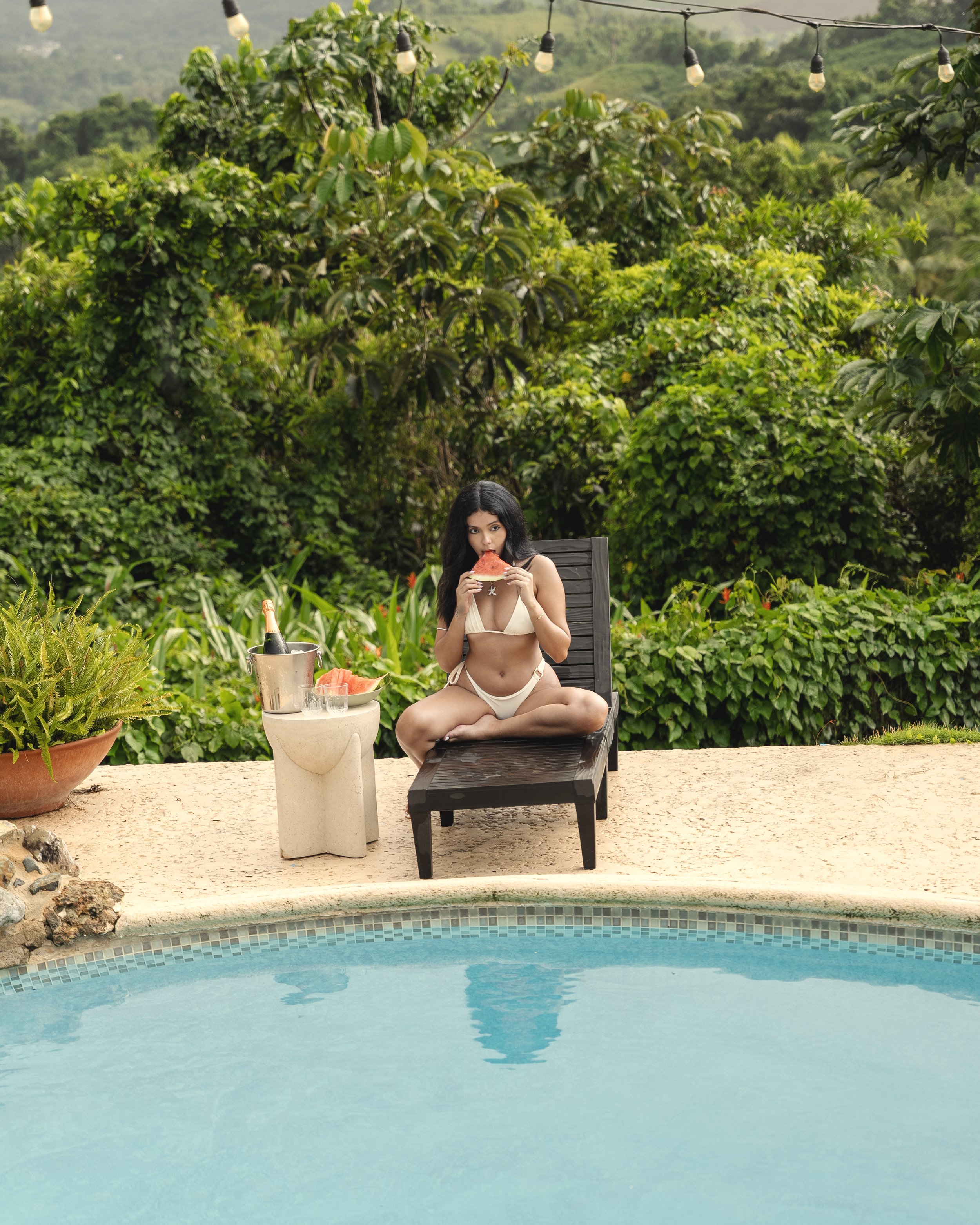 A woman in a beige bikini is sitting cross-legged on a lounge chair by a swimming pool, eating a slice of watermelon. There is a small round table next to her with a bucket of champagne, a sliced watermelon, and two empty glasses. The background features lush green vegetation and outdoor string lights hanging overhead.