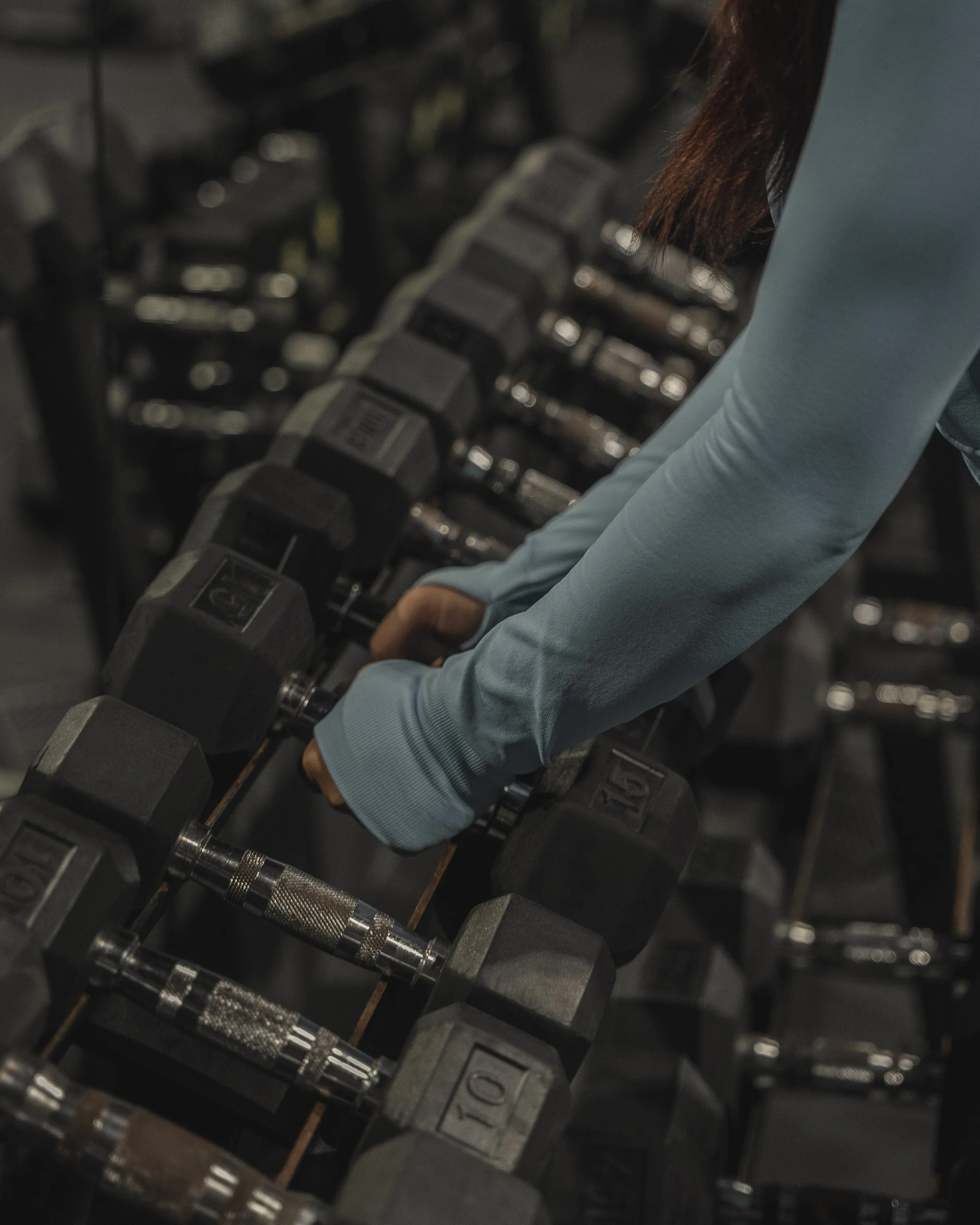 A person with long hair and a light blue long sleeve shirt adjusting a dumbbell on a rack in a gym.