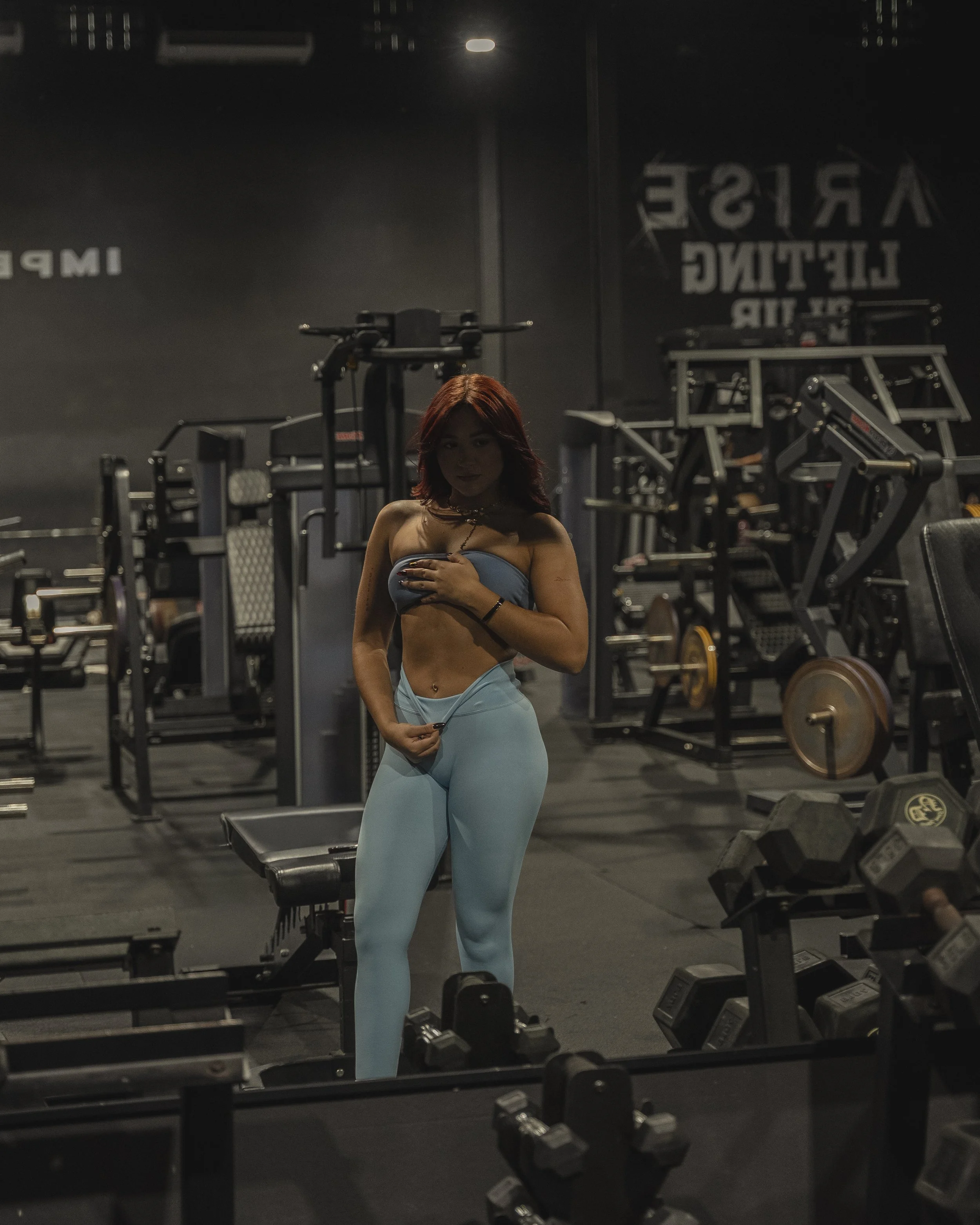 A woman with auburn hair wearing light blue workout leggings and a strapless top, holding a small weight close to her chest, standing in a gym in front of a mirror among workout equipment, with the text 'ARISE LIGHTING' reflected in the mirror.