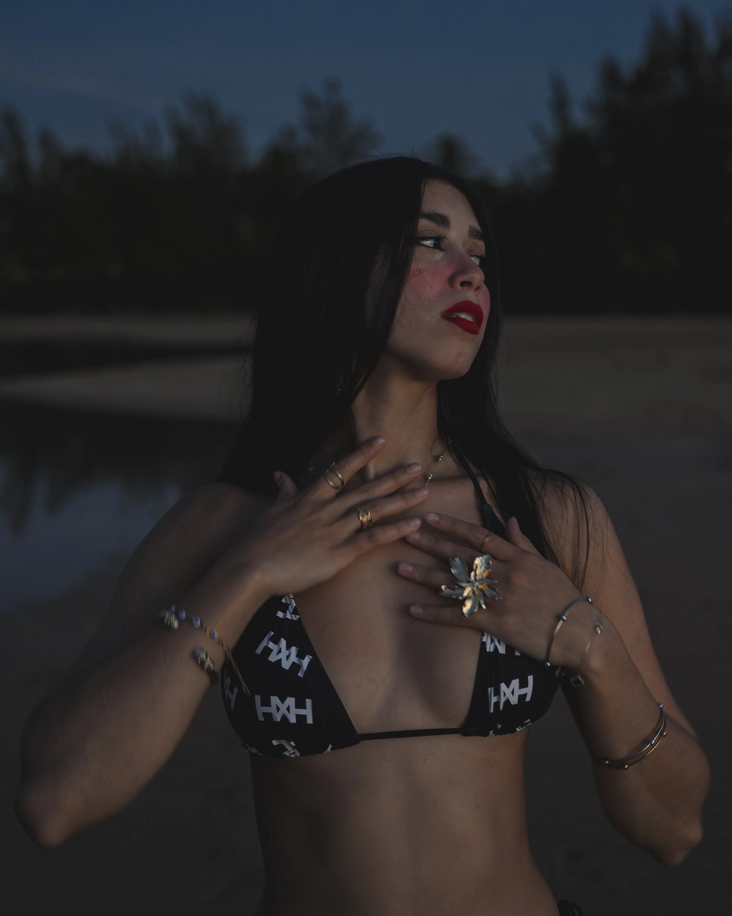 A woman wearing a black crop top with white lettering stands outdoors during twilight. She has dark hair, red lipstick, and adorned with gold jewelry, including a large butterfly necklace and multiple rings and bracelets. She is touching her chest and has a contemplative expression.