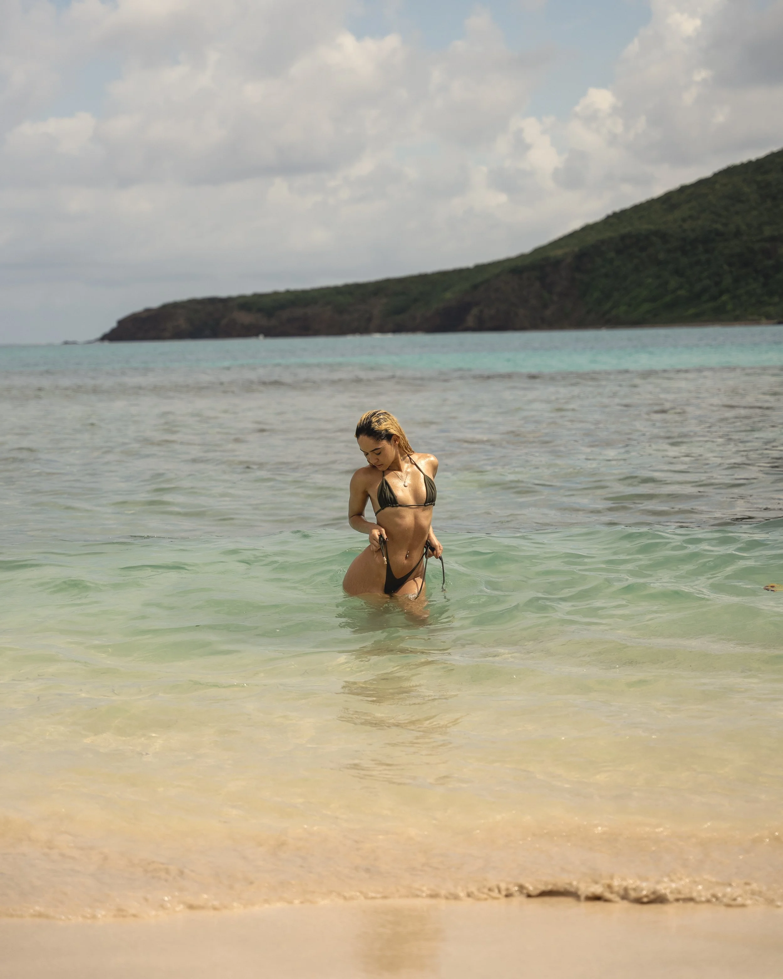 A woman with wet hair in a black bikini standing in the ocean near the shore, with a green hillside and a cloudy sky in the background.