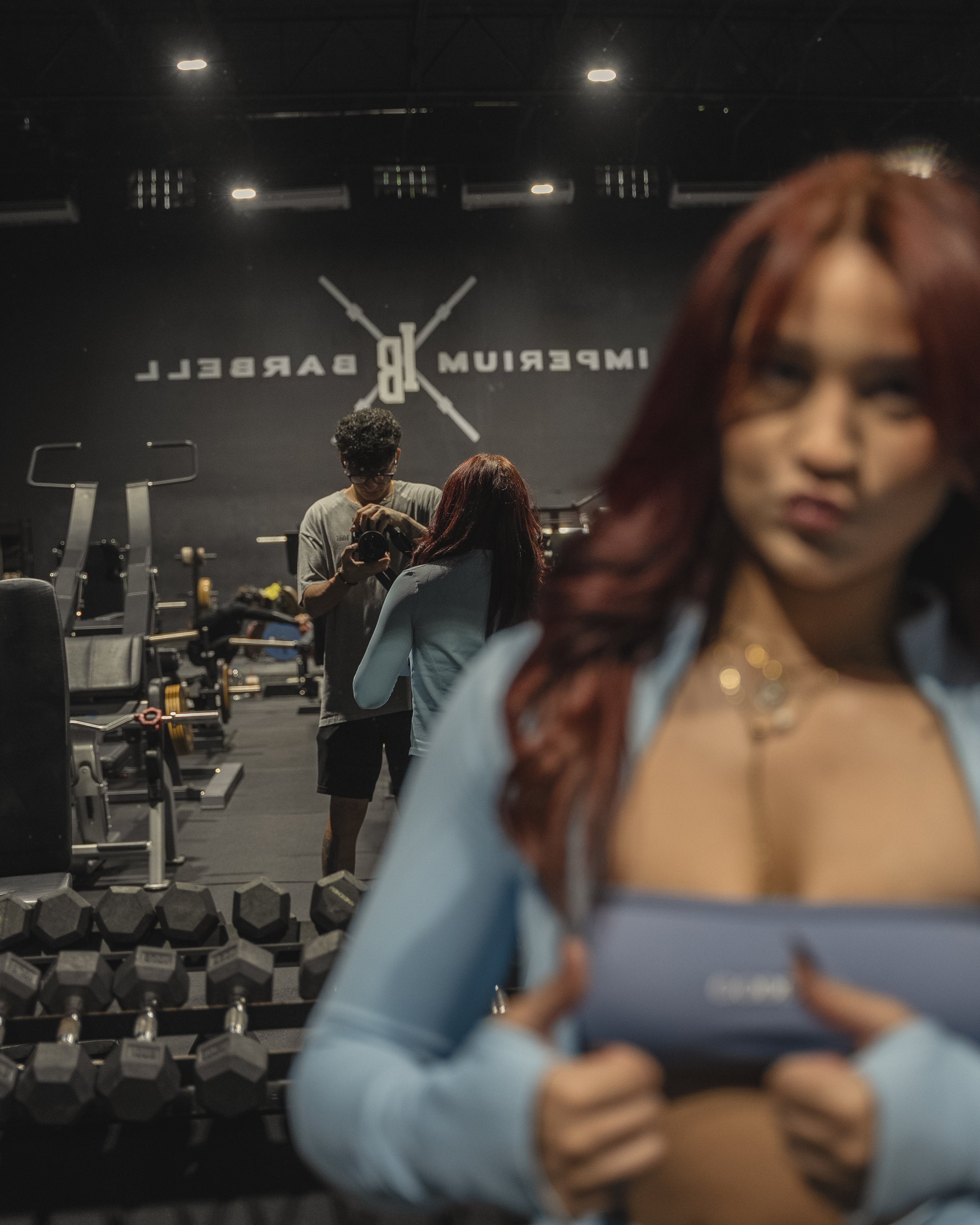 Mirror selfie in a gym with three people, weightlifting equipment including dumbbells, and a sign that reads 'Impressive Barrel' on the wall.