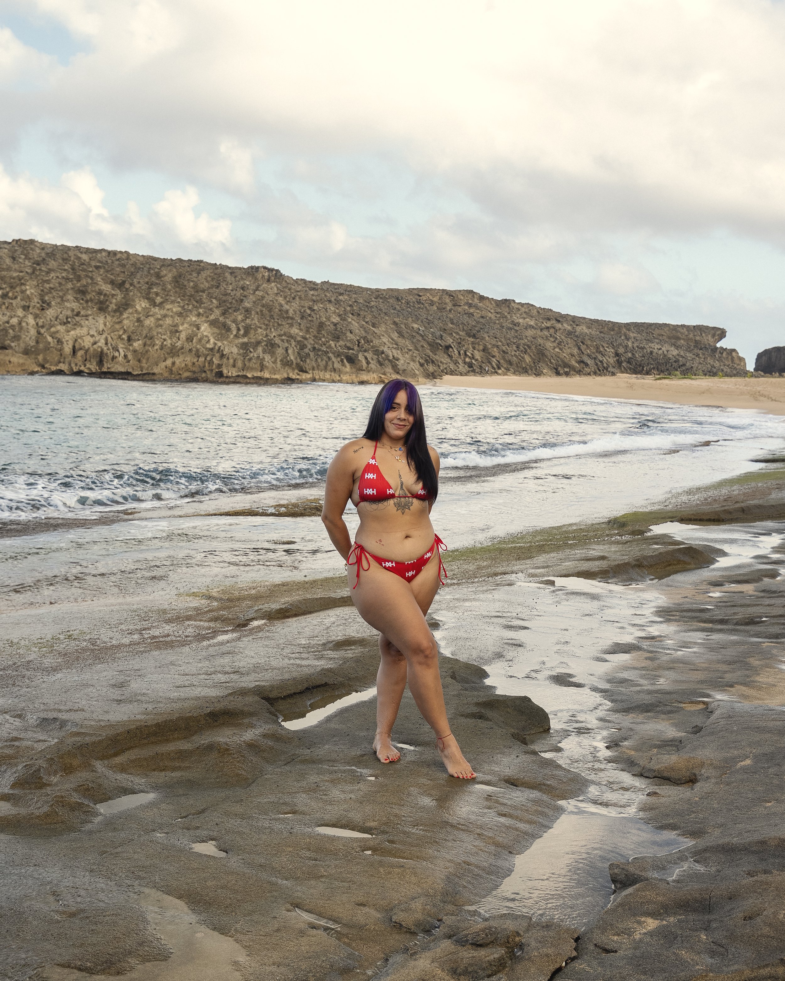 A woman in a red bikini with white patterns stands on a rocky beach near the ocean, with cliffs in the background and partly cloudy skies.
