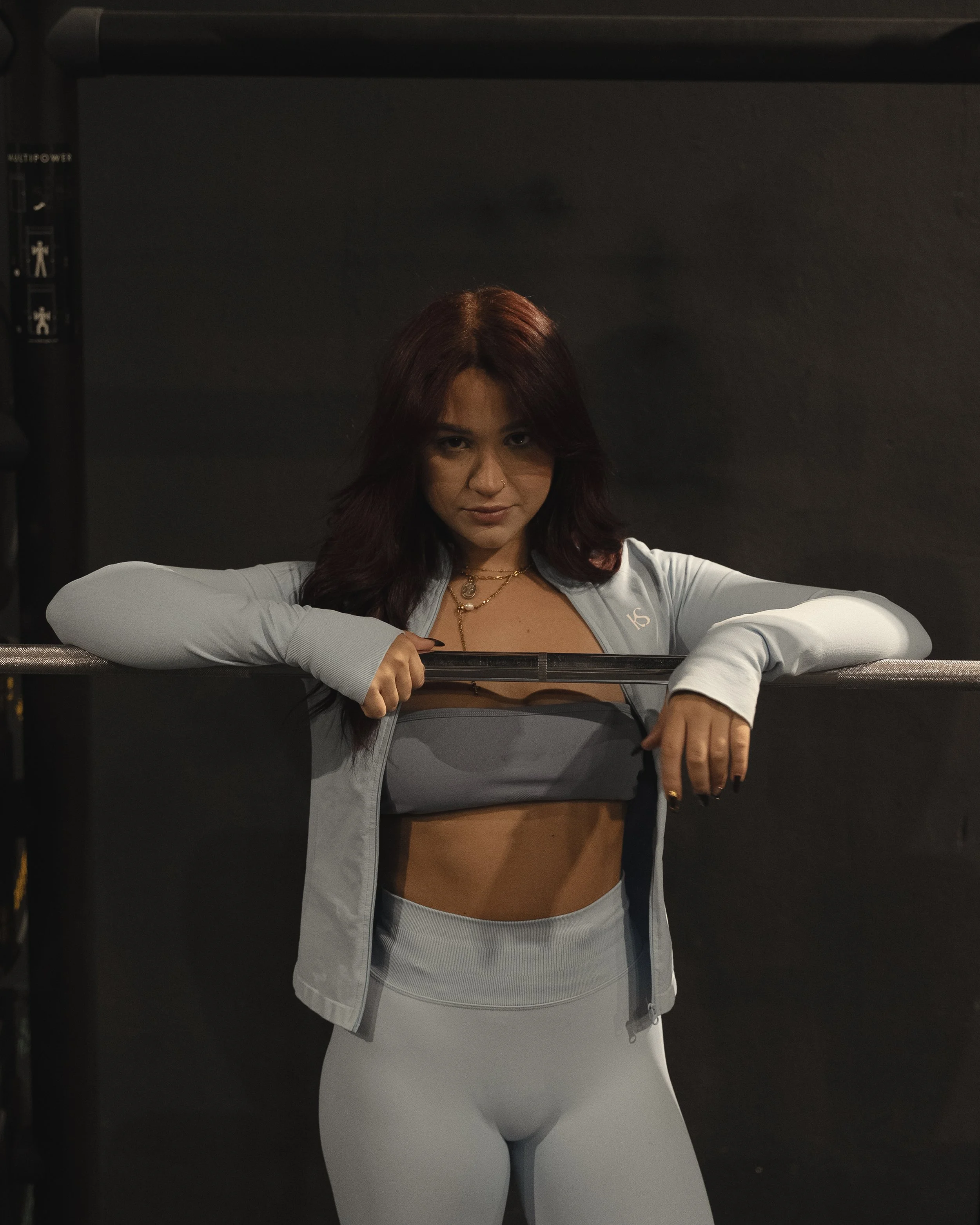 Young woman with reddish-brown hair wearing a grey jacket and white leggings, seated at a makeup mirror with her arms resting on a black bar, in a dimly lit room with a dark background.