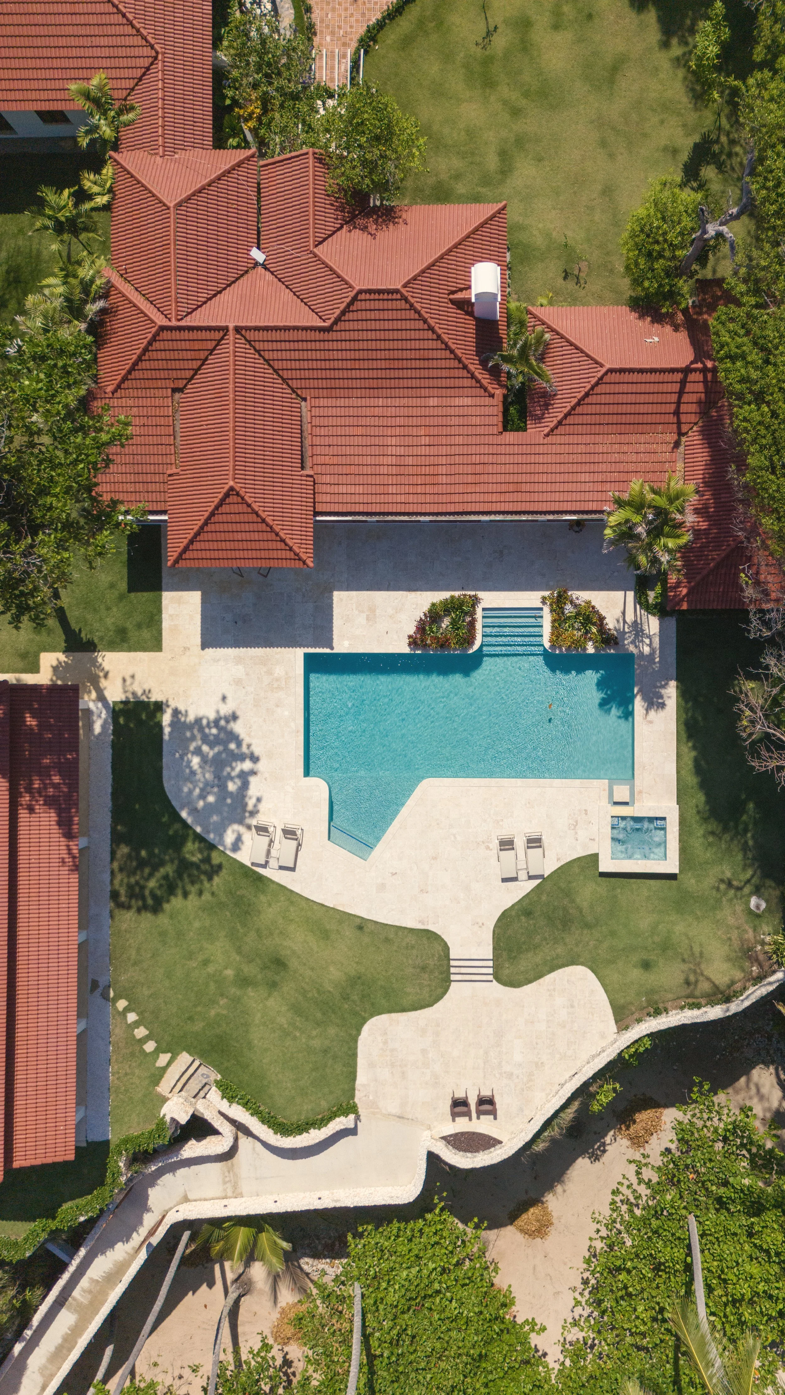 Aerial view of a house with a red-tiled roof, a swimming pool, surrounding greenery, palm trees, and a landscaped yard.