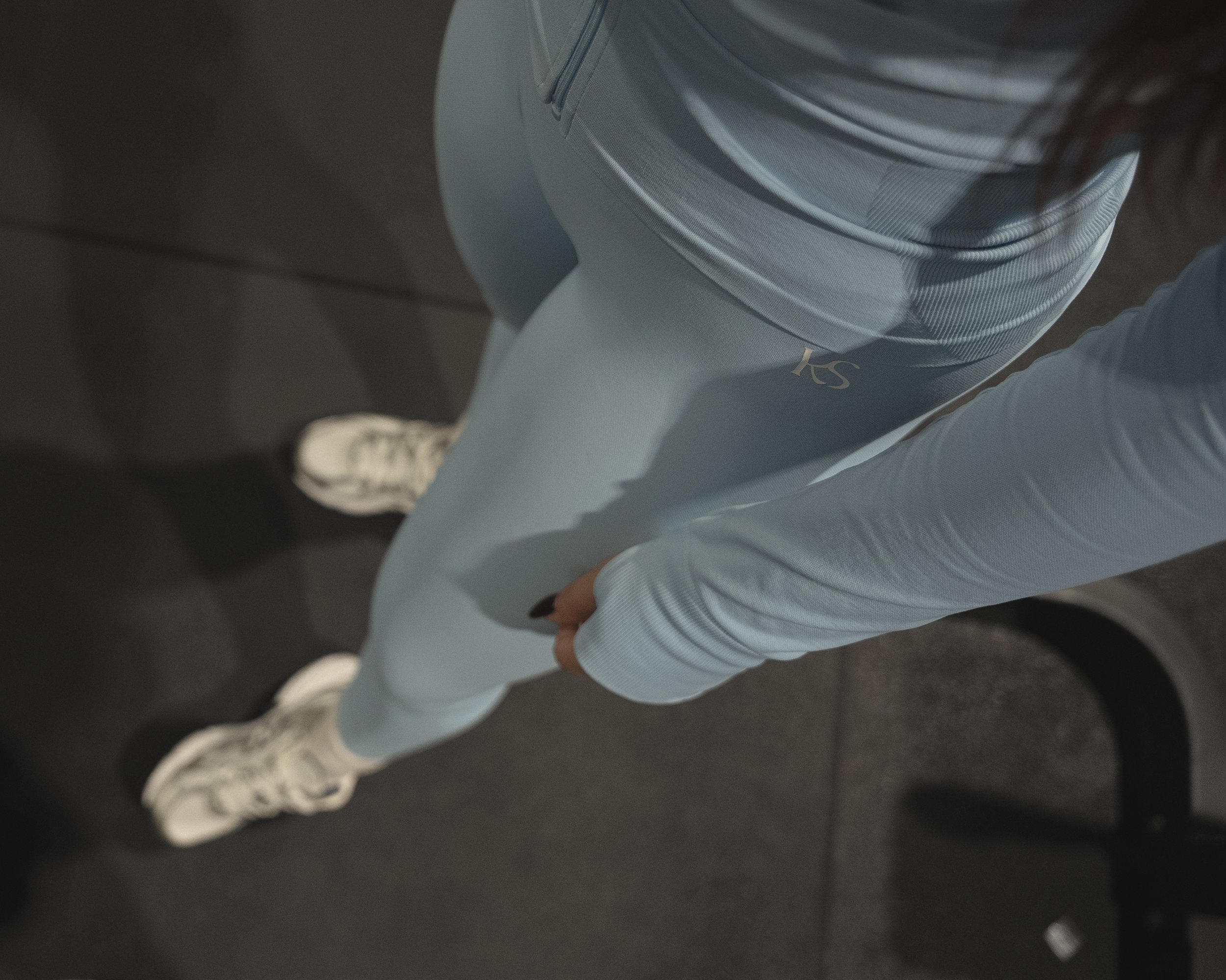 Close-up of a person wearing light blue pants with the word 'KSS' on the left thigh, standing on a dark floor with white sneakers visible.