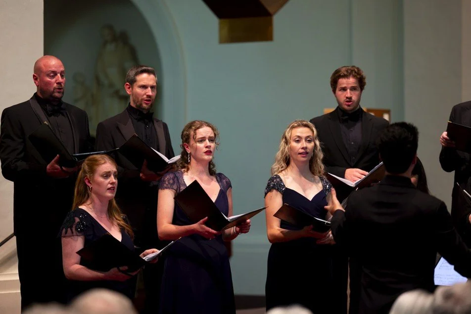 A choir and conductor performing on a stage inside a church or concert hall. The stage has candelabras, purple banners, and a piano on the left. Audience members are seated in front, some holding music sheets and devices.