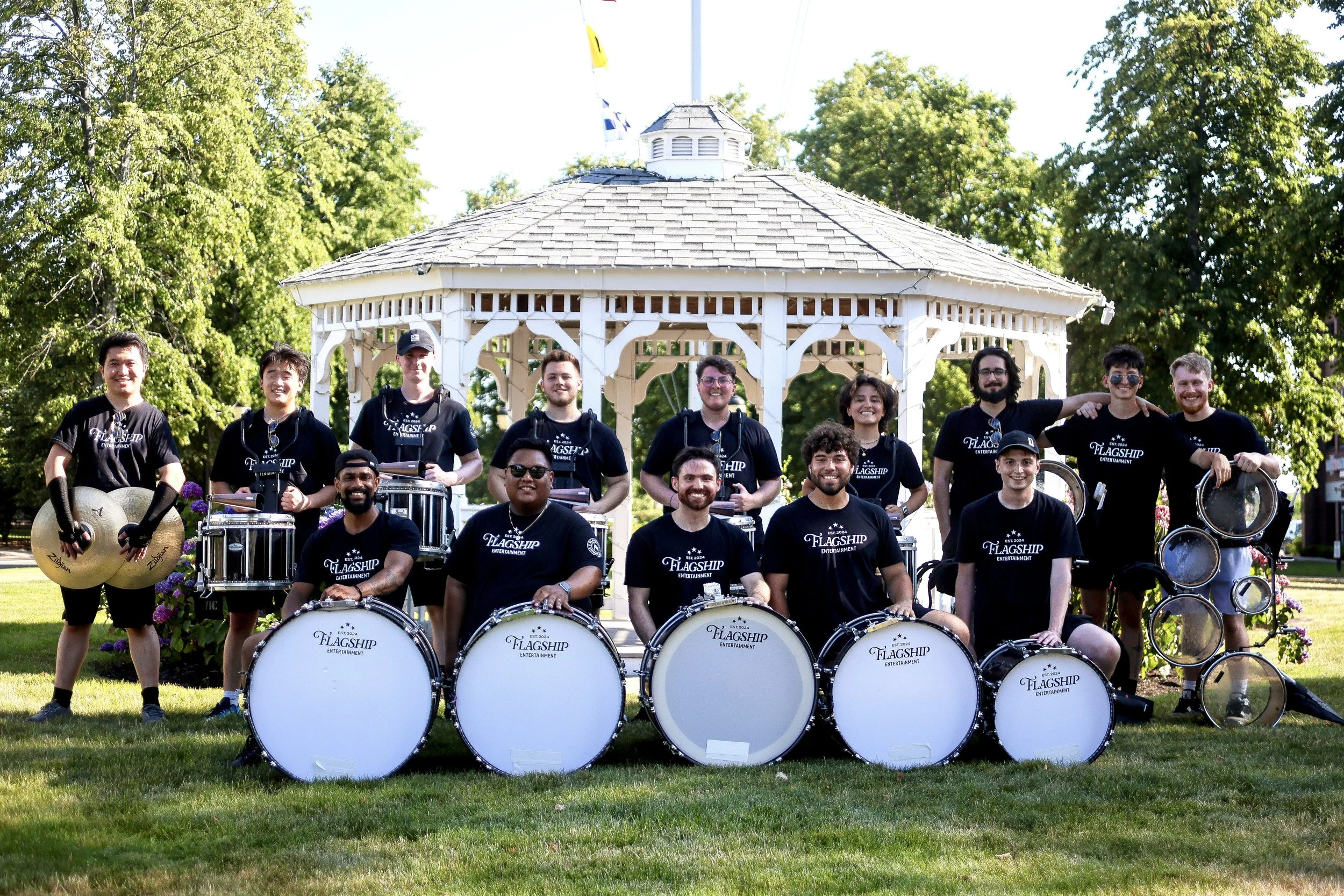 A group of people in black T-shirts with 'Flagship Entertainment' logo, posing with musical instruments in front of a white gazebo in a park with green trees.