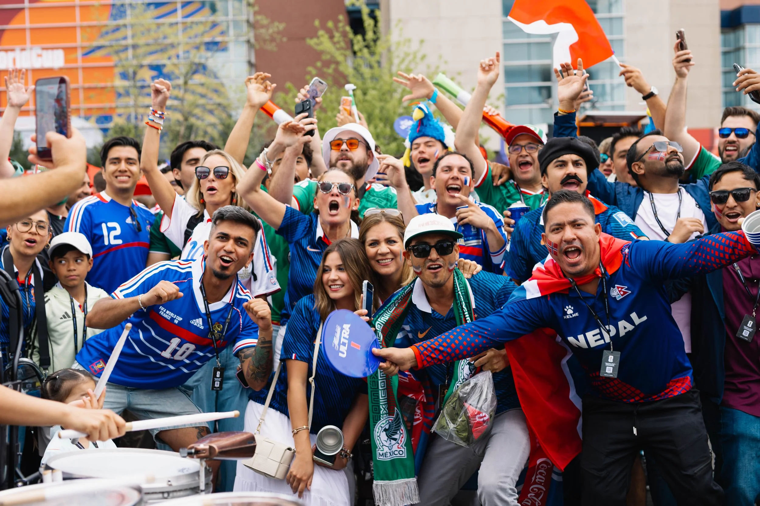 A large group of people celebrating outdoors, waving flags and taking selfies. Many are wearing soccer jerseys, face paint, and accessories supporting their countries.