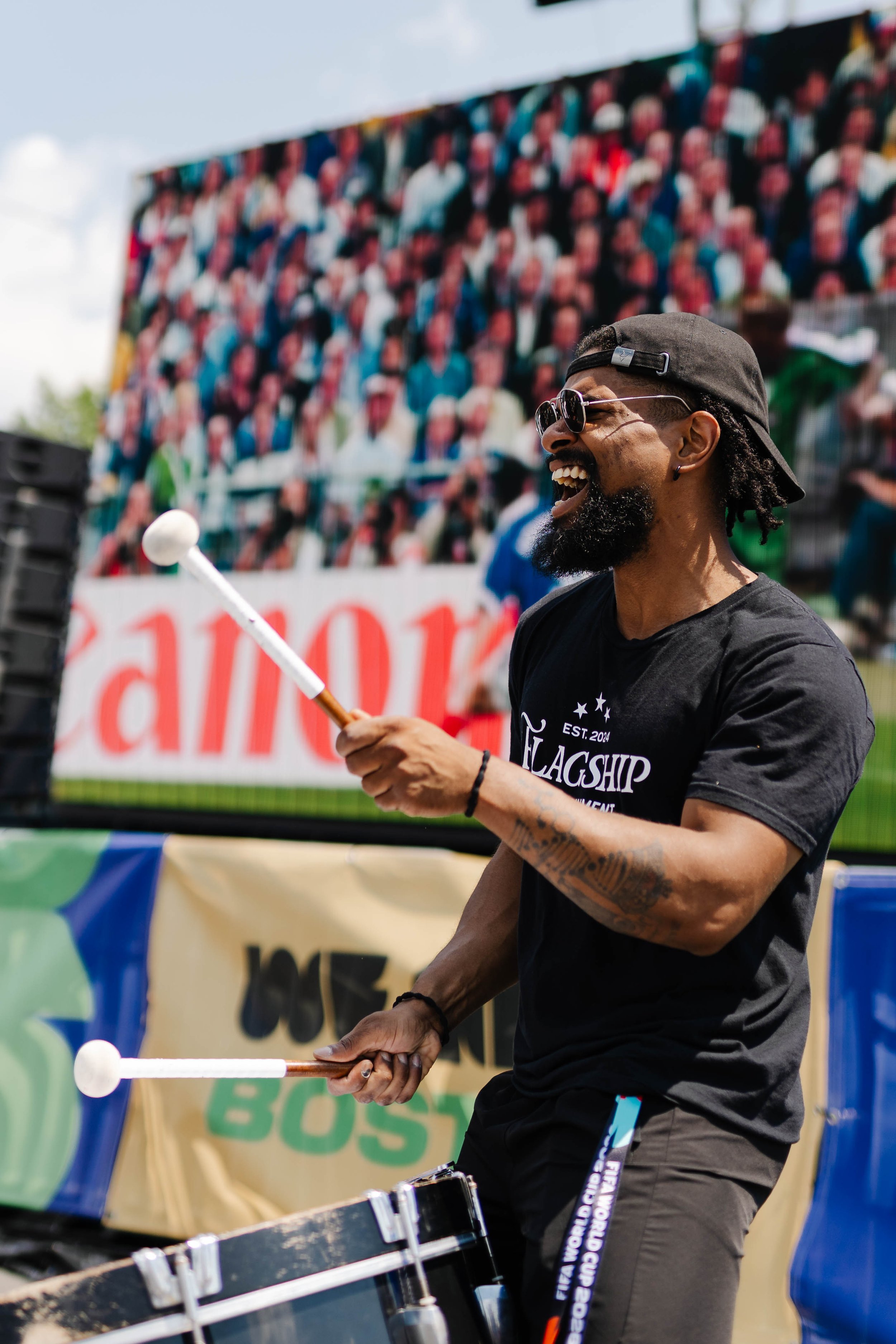 A man playing drums at an outdoor event, smiling and wearing sunglasses, a black t-shirt, and a black cap backwards. In the background is a colorful crowd mural.