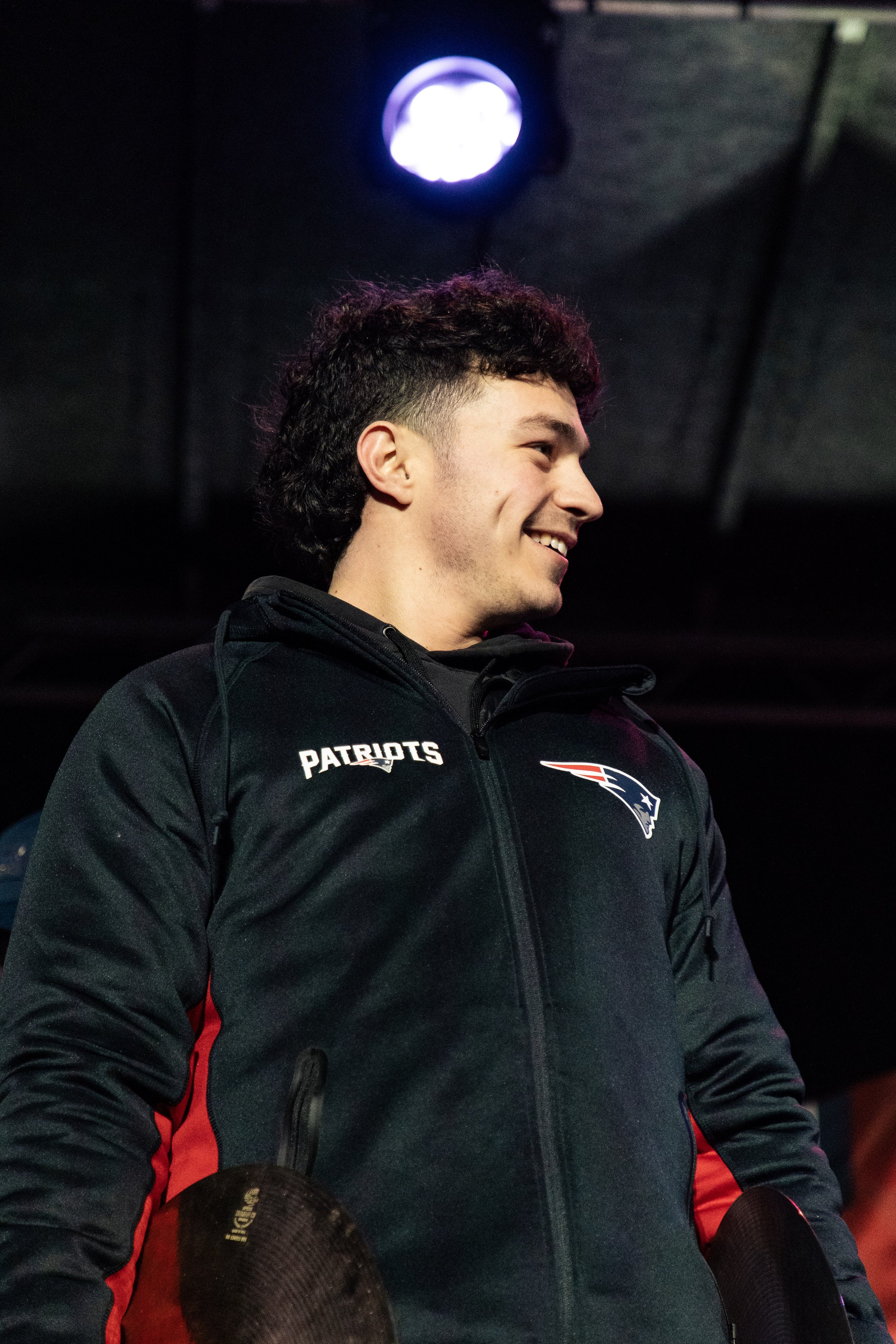 Young man wearing a black Patriots jacket, smiling, holding a pair of black and red football gloves, standing indoors with a dark background and a purple and white light above his head.