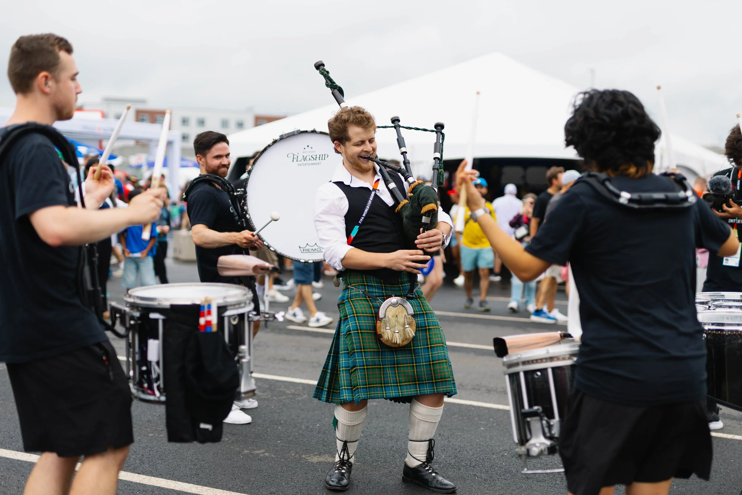 Musicians playing drums while a man in traditional Scottish attire dances at an outdoor event.