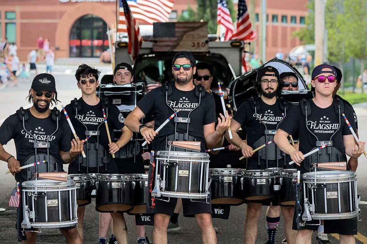 Group of men playing drums in a parade, wearing black shirts with 'Flagship Entertainment' logo, with American flags in the background