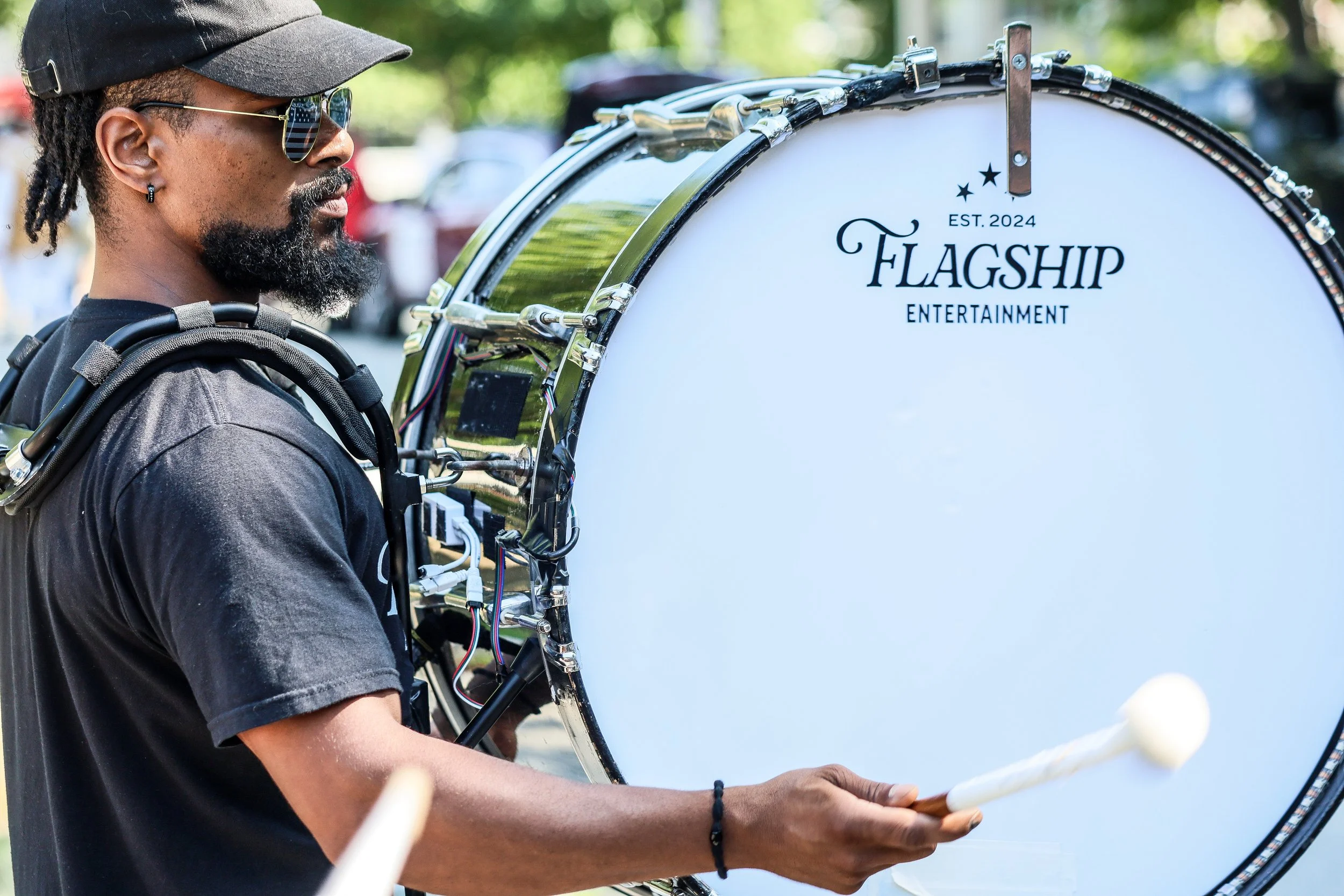 A man with a beard and dreadlocks, wearing sunglasses and a black cap, playing a bass drum outdoors. The drum has a white head with the words 'Flagship Entertainment' and 'Est. 2024' printed on it.