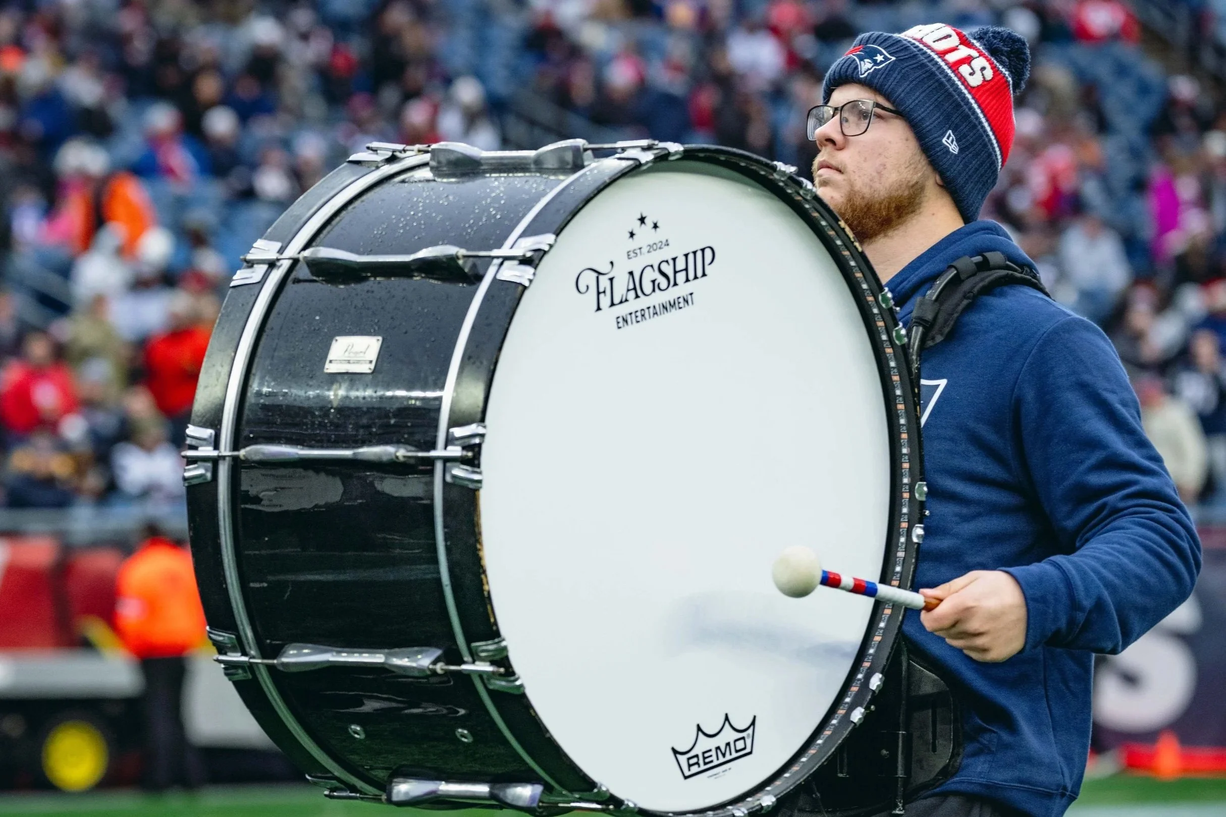 A man wearing glasses, a knit cap, and a hoodie is playing a bass drum at a sports stadium. The drum is black and white with the words 'Flagship Entertainment' and 'EST. 2024' printed on it. The background shows a crowd of spectators at Gillette.