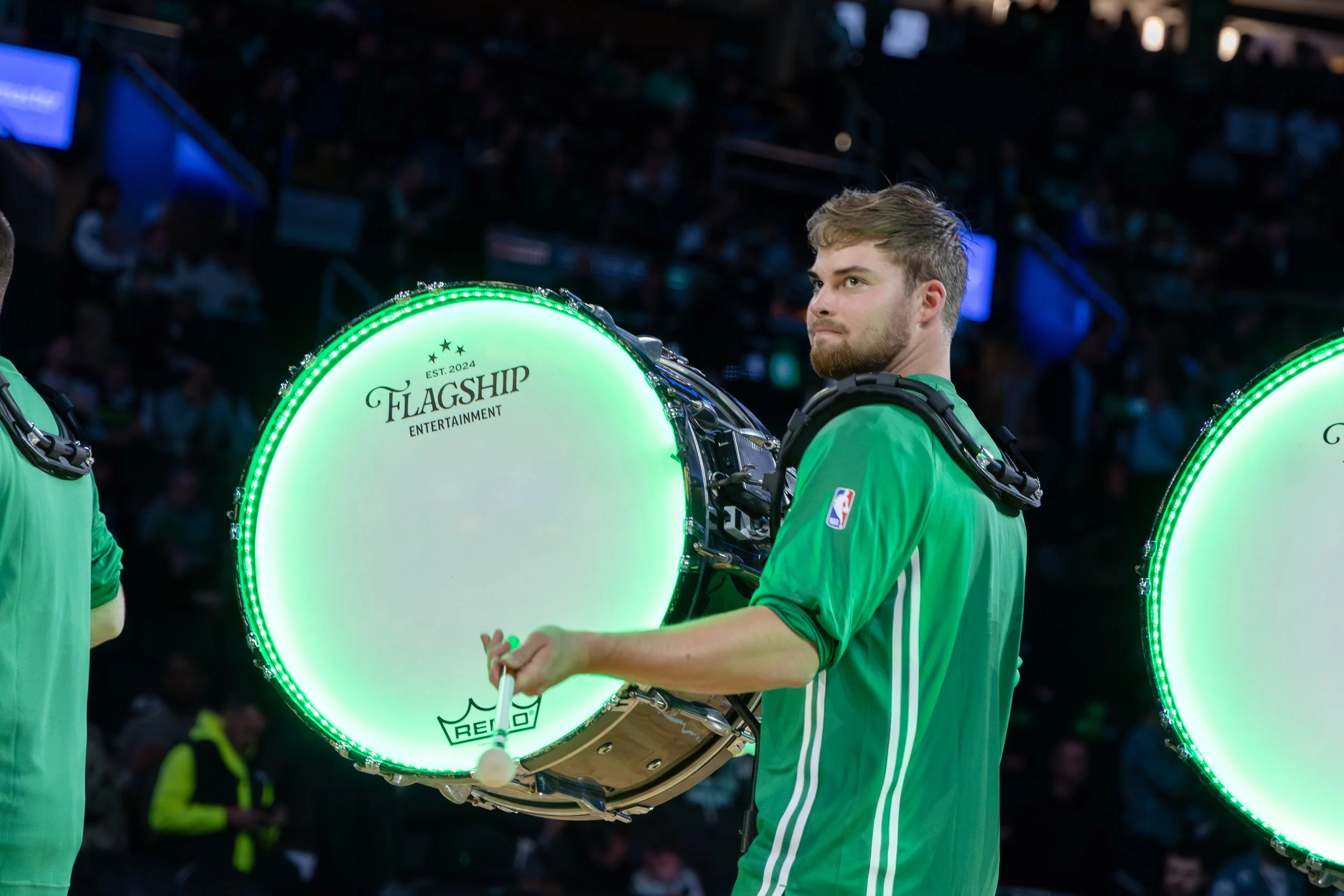 A young man with a beard and short, styled hair wearing a green sports jersey with an NBA logo on the sleeve, standing in a dimly lit arena, playing a bass drum with a glowing white rim and black logo that reads 'Flagship Entertainment,' while anothe