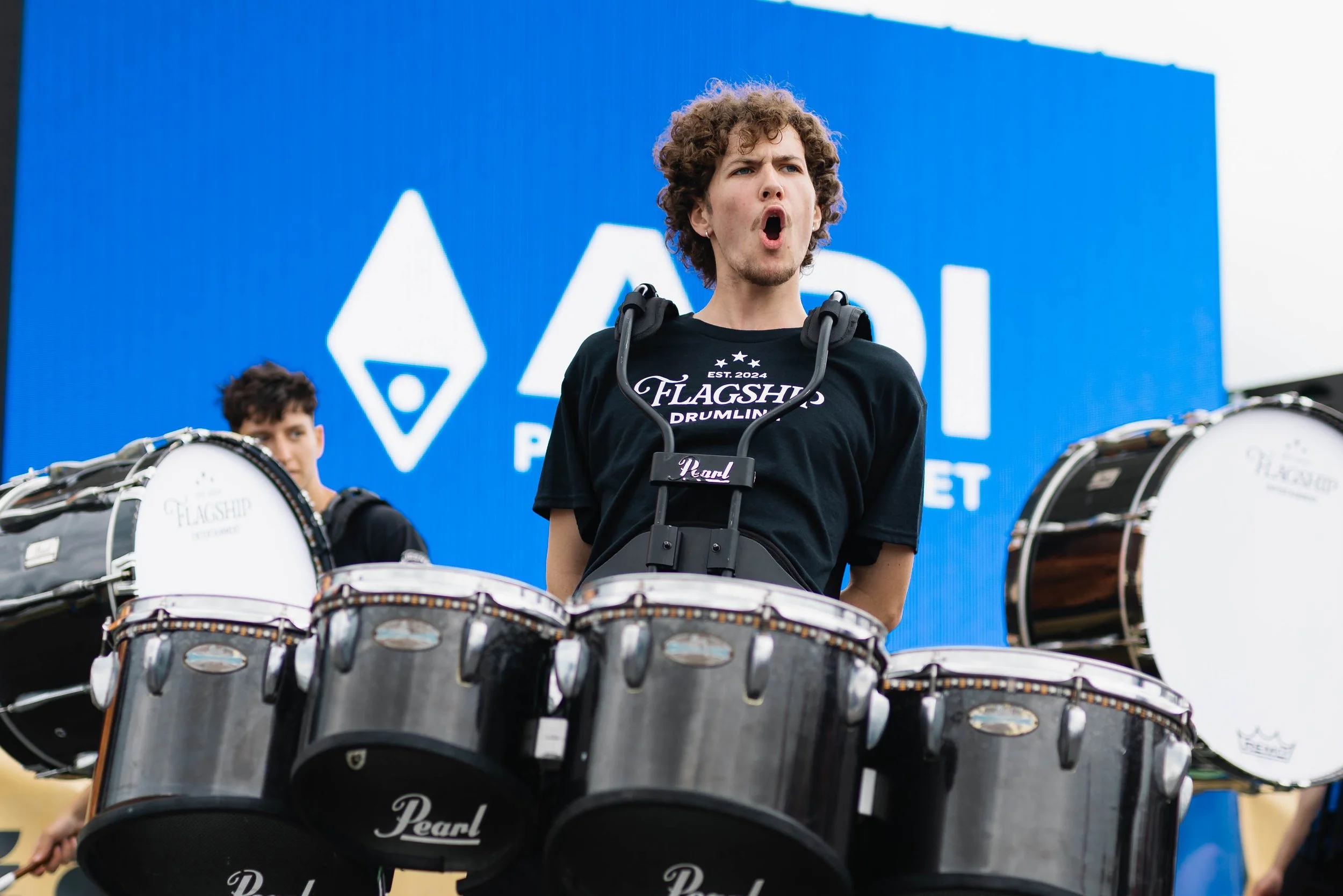 Young man with curly hair playing drums outdoors, wearing a black T-shirt with 'Flagship Drumming' logo, with blue background and drums in foreground.