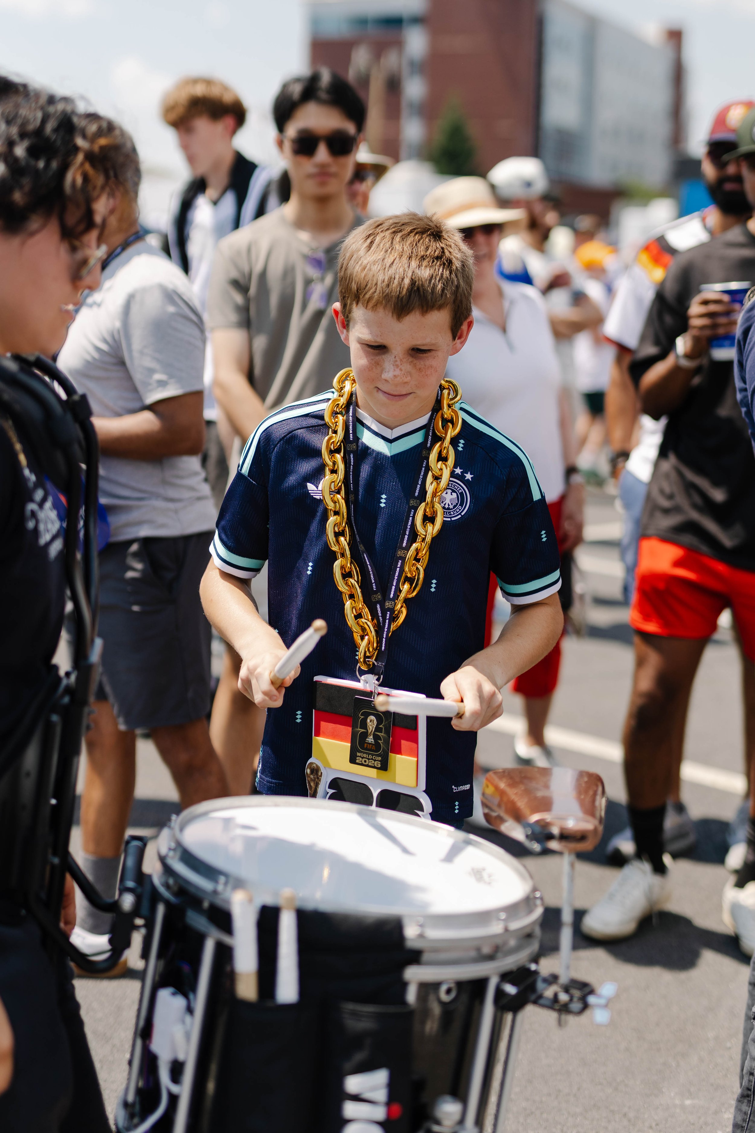 A young boy with a German national football team jersey is wearing a large gold chain and holding a small FIFA World Cup 2026-themed bag, standing among other people in an outdoor setting.