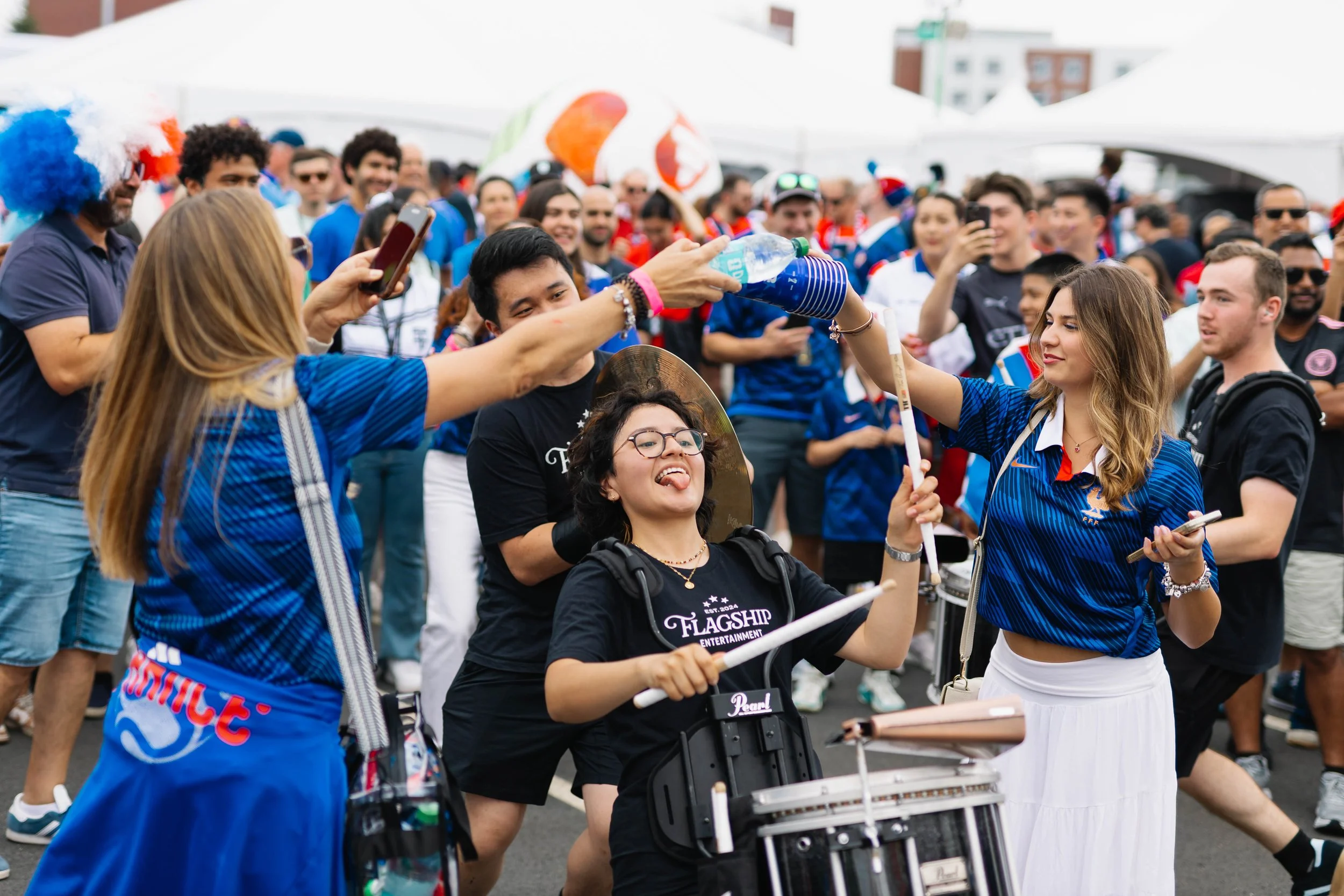 Crowd of people at an outdoor event, some wearing sports jerseys, with a person playing drums in the foreground.