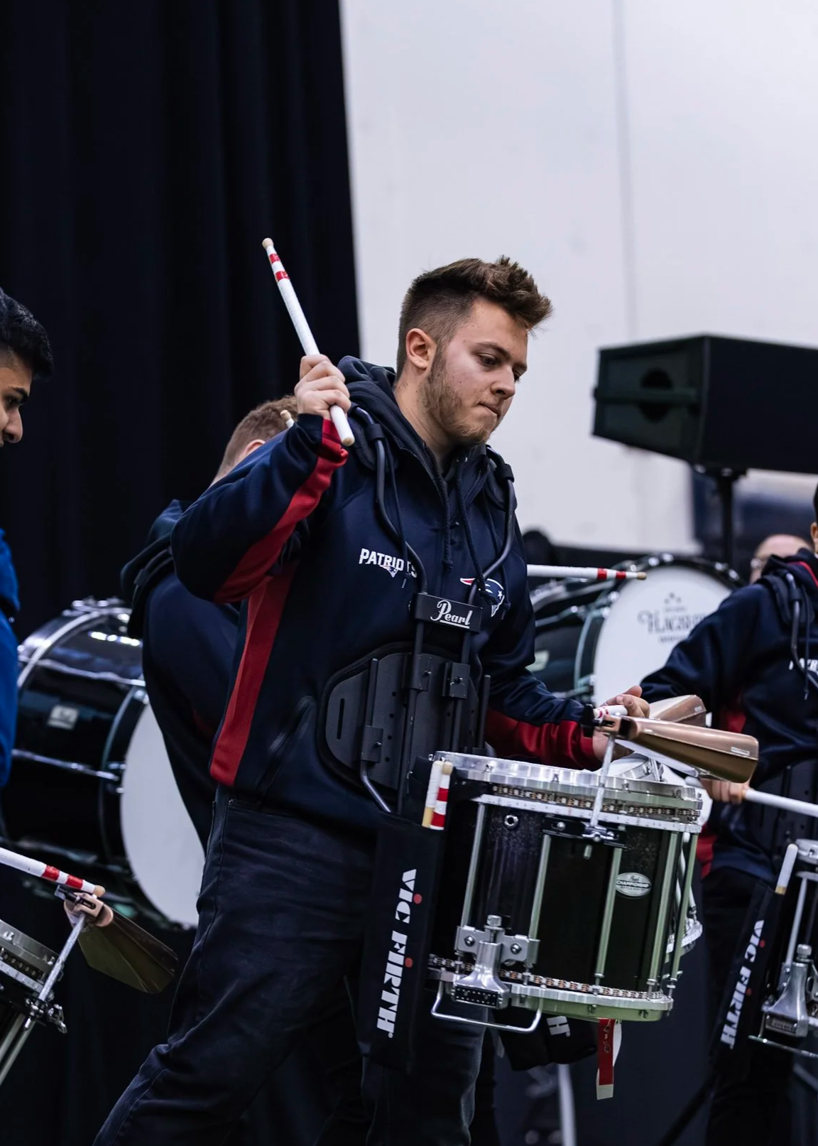 Young male drummer with a beard and short hair playing drums at a concert, wearing a black t-shirt with white text and a lanyard with a badge that says 'Pearl' around his neck.