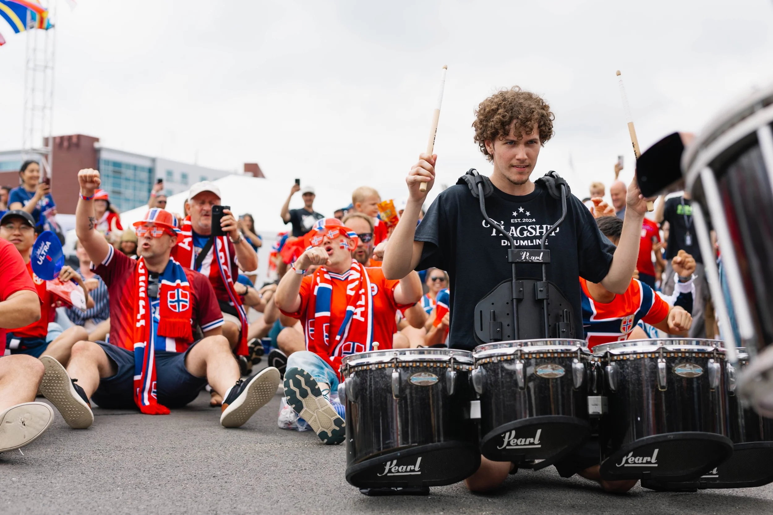 Young man drumming at a crowded outdoor event, surrounded by fans wearing Norwegian flags and scarves, with people taking photos and videos.